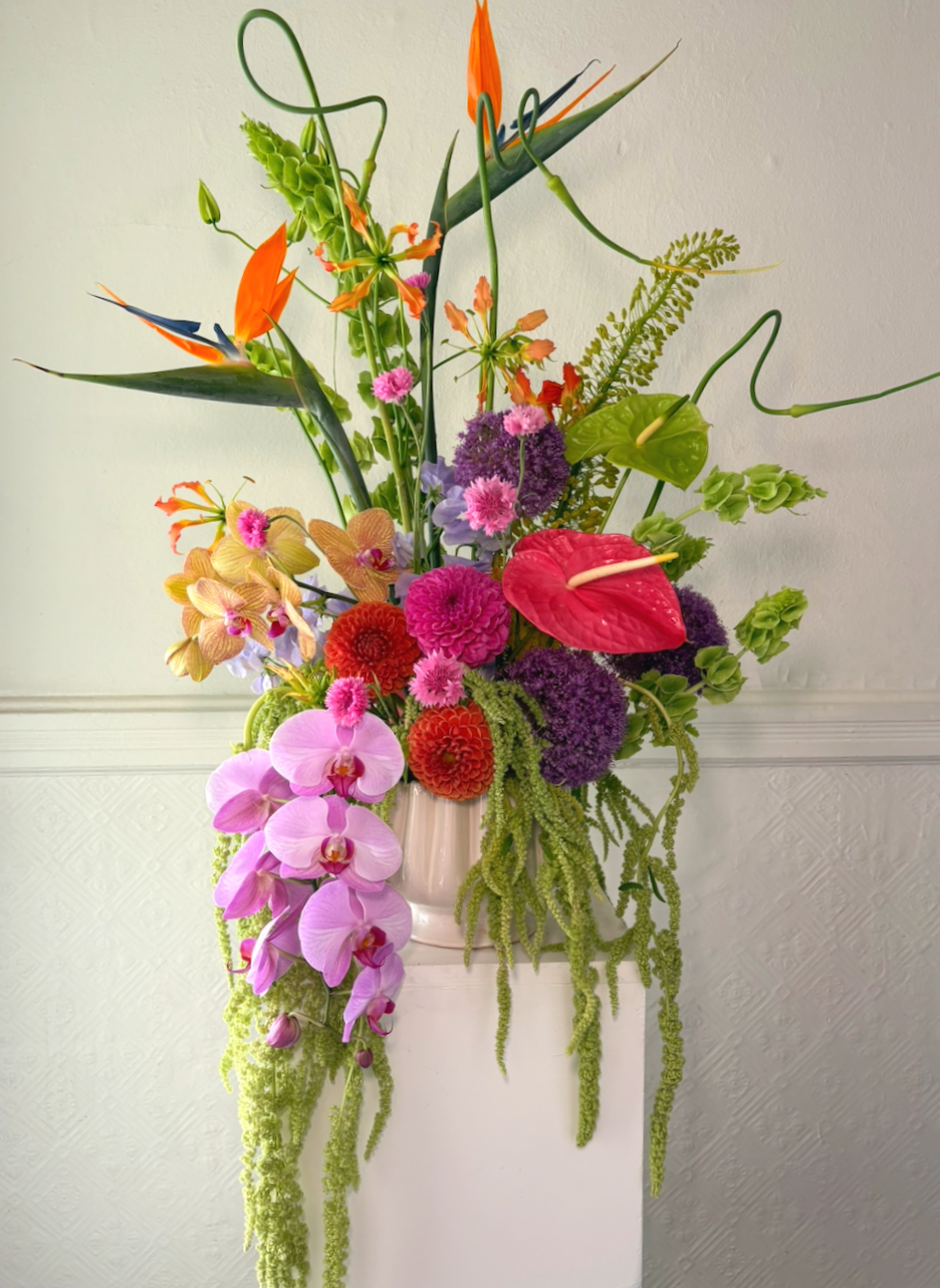 A tall, colorful floral arrangement in a white vase featuring orchids, roses, anthuriums, and various greenery and tropical flowers