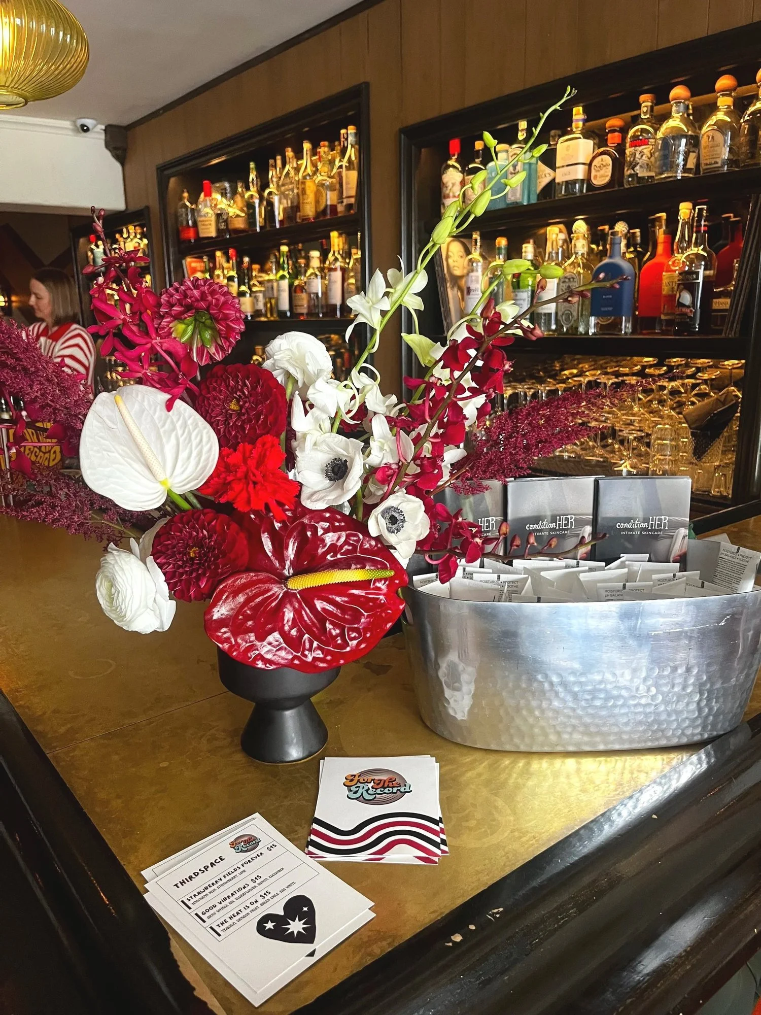 A table with a vibrant floral arrangement in a black vase, a metal bucket filled with small packages labeled 'condition HER,' and flyers placed on the table. In the background, a bar with shelves of various liquor bottles is visible, and only one person, a woman in a red and white striped shirt, can be seen.