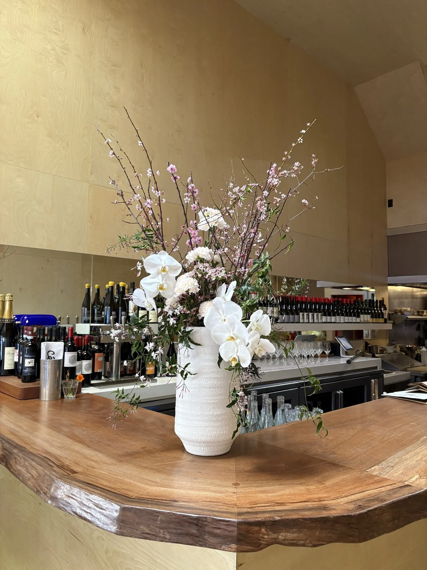 A white vase with a flower arrangement of pink and white blossoms and green. leaves on a wooden bar counter in a restaurant or bar setting.