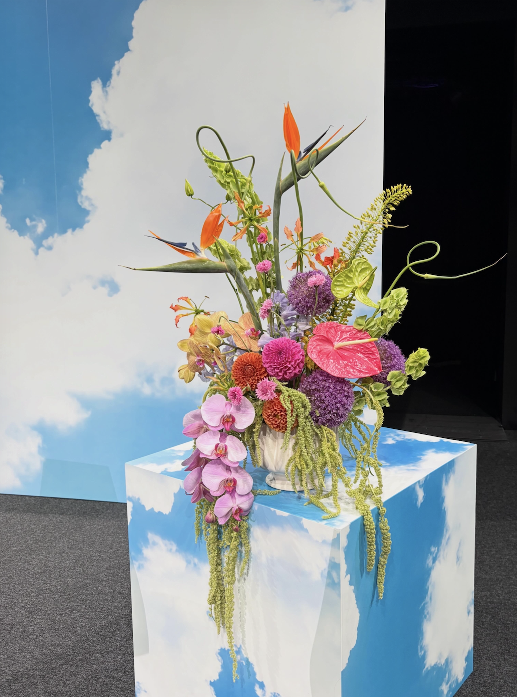 Colorful flower arrangement with pink, purple, orange, and green flowers in a vase on a sky-themed pedestal, with blue sky and white clouds in the background.