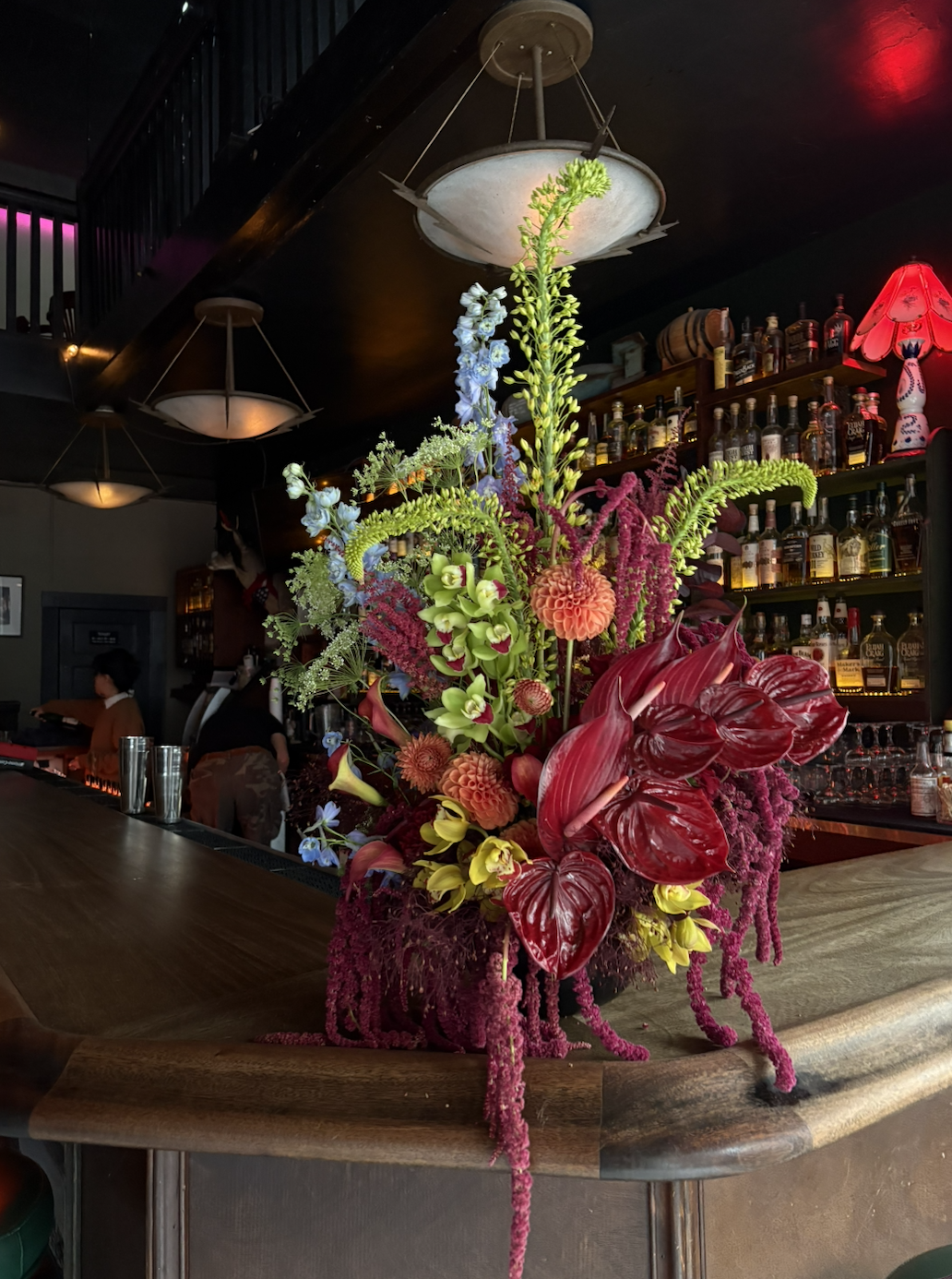 A large floral arrangement with red, purple, green, and blue flowers on a wooden bar in a dimly lit bar or restaurant.