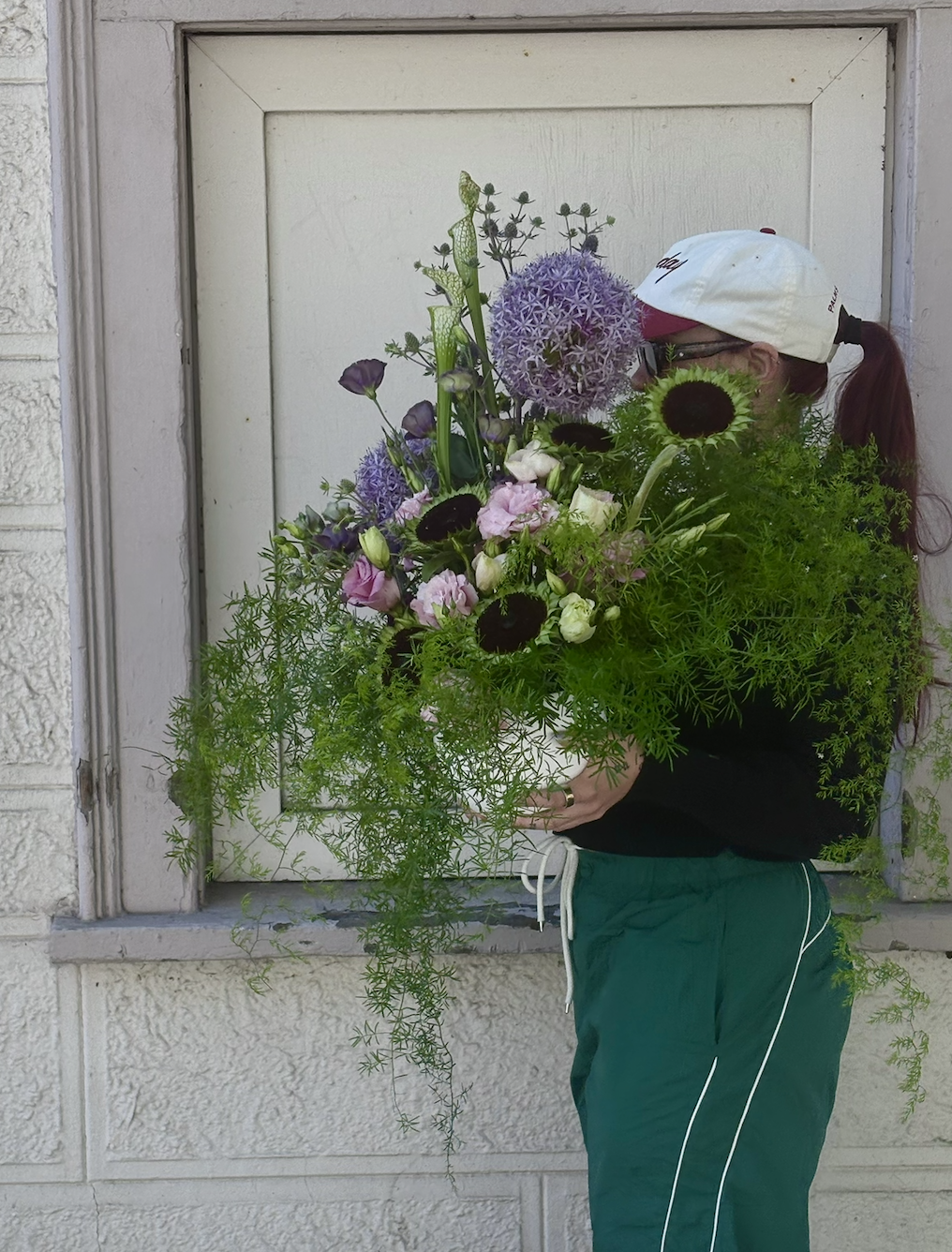 Person with long hair in a ponytail, wearing a white cap, glasses, a black sweatshirt, and green pants, is holding a large colorful flower arrangement in front of a closed white window.