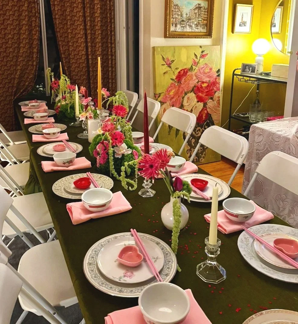 A dining table set for a celebration with pink, white, and red floral decorations, candles, and dinnerware, surrounded by white chairs in a decorated room with paintings and artwork on the walls.