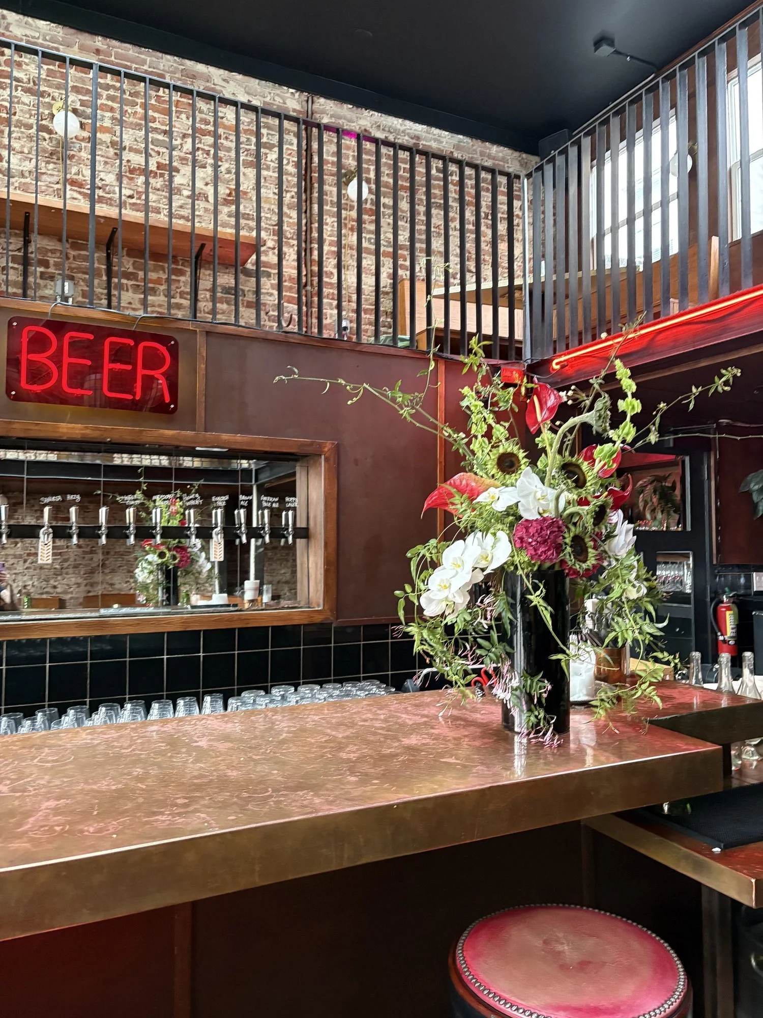 A bar area with a bouquet of flowers on a marble countertop, neon 'BEER' sign, exposed brick wall, black tile accents, and a two-story layout with a wooden railing and modern lighting.