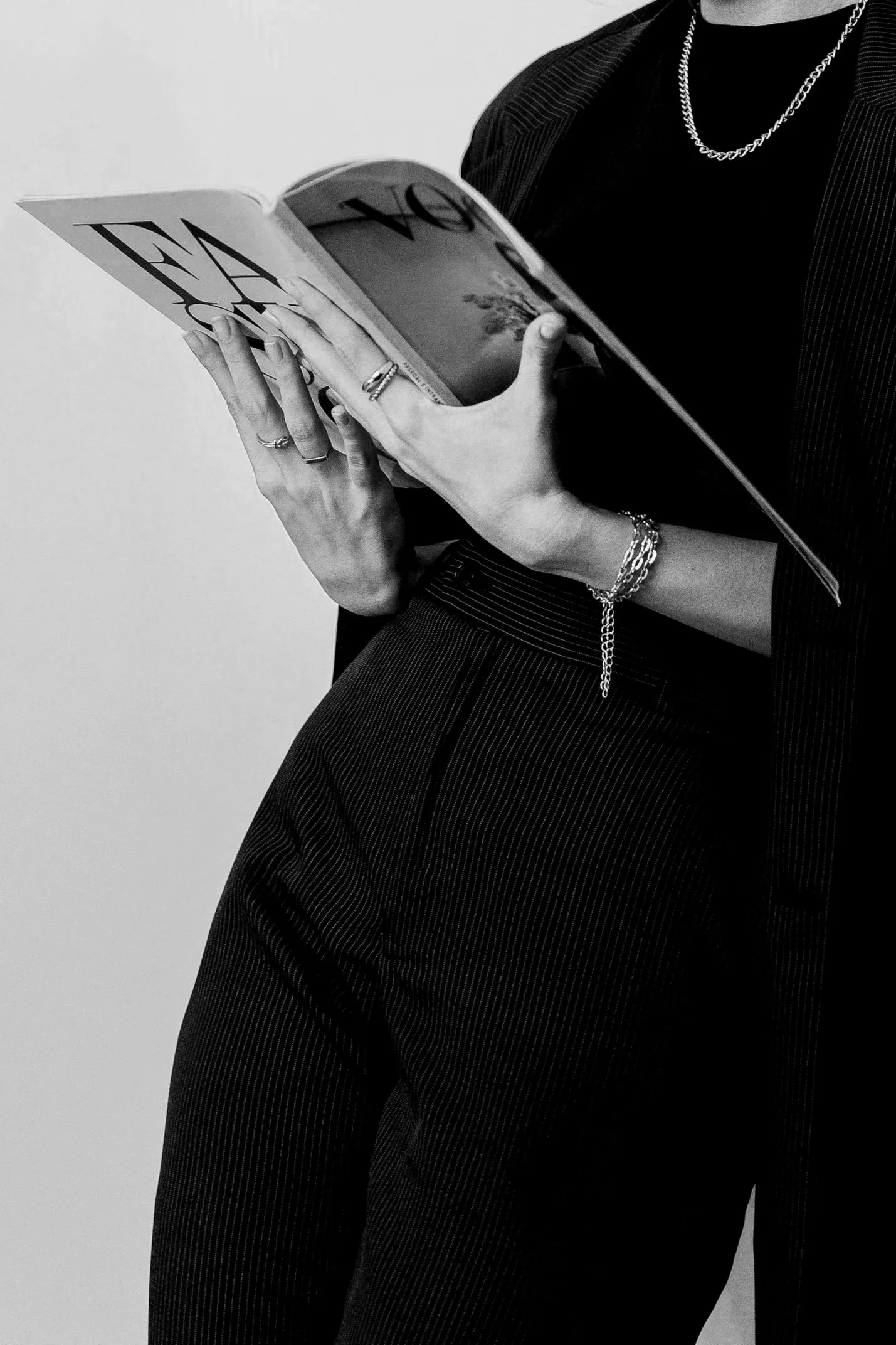 A person dressed in a pinstriped suit, holding an open magazine with both hands, wearing rings, a bracelet, a chain necklace, and a watch. The image is in black and white.