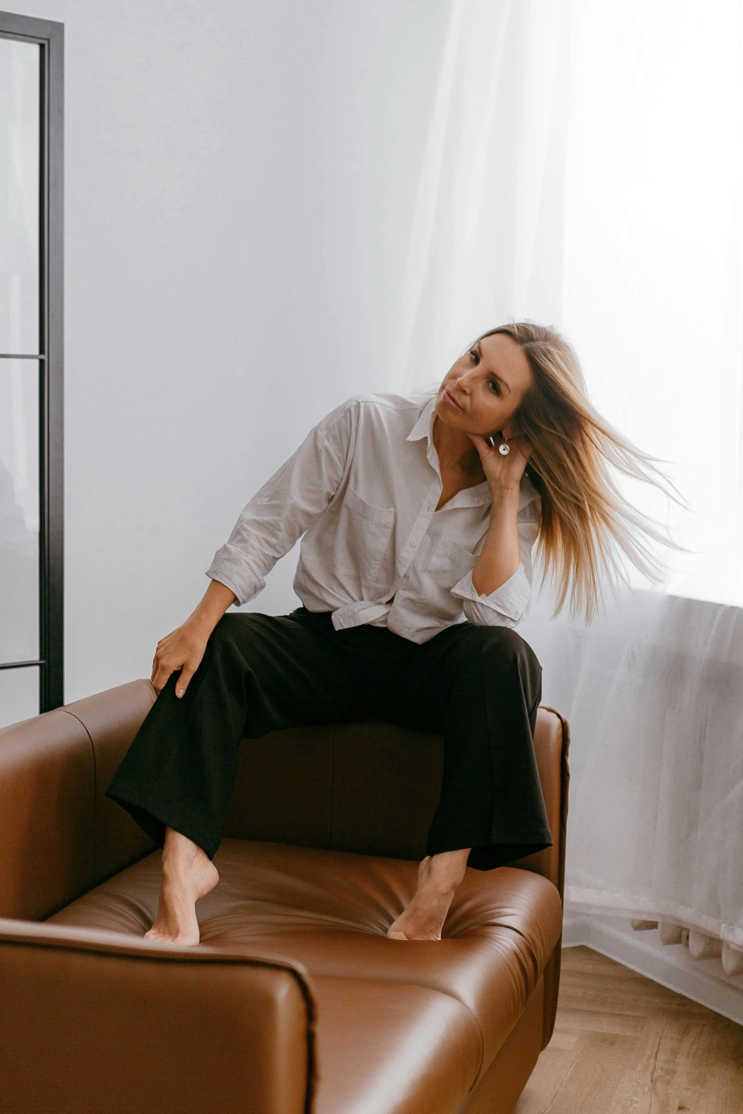 A woman with blonde hair, wearing a white shirt and black pants, sitting on a leather couch with her feet on the cushions, looking contemplative in a bright room with white curtains.