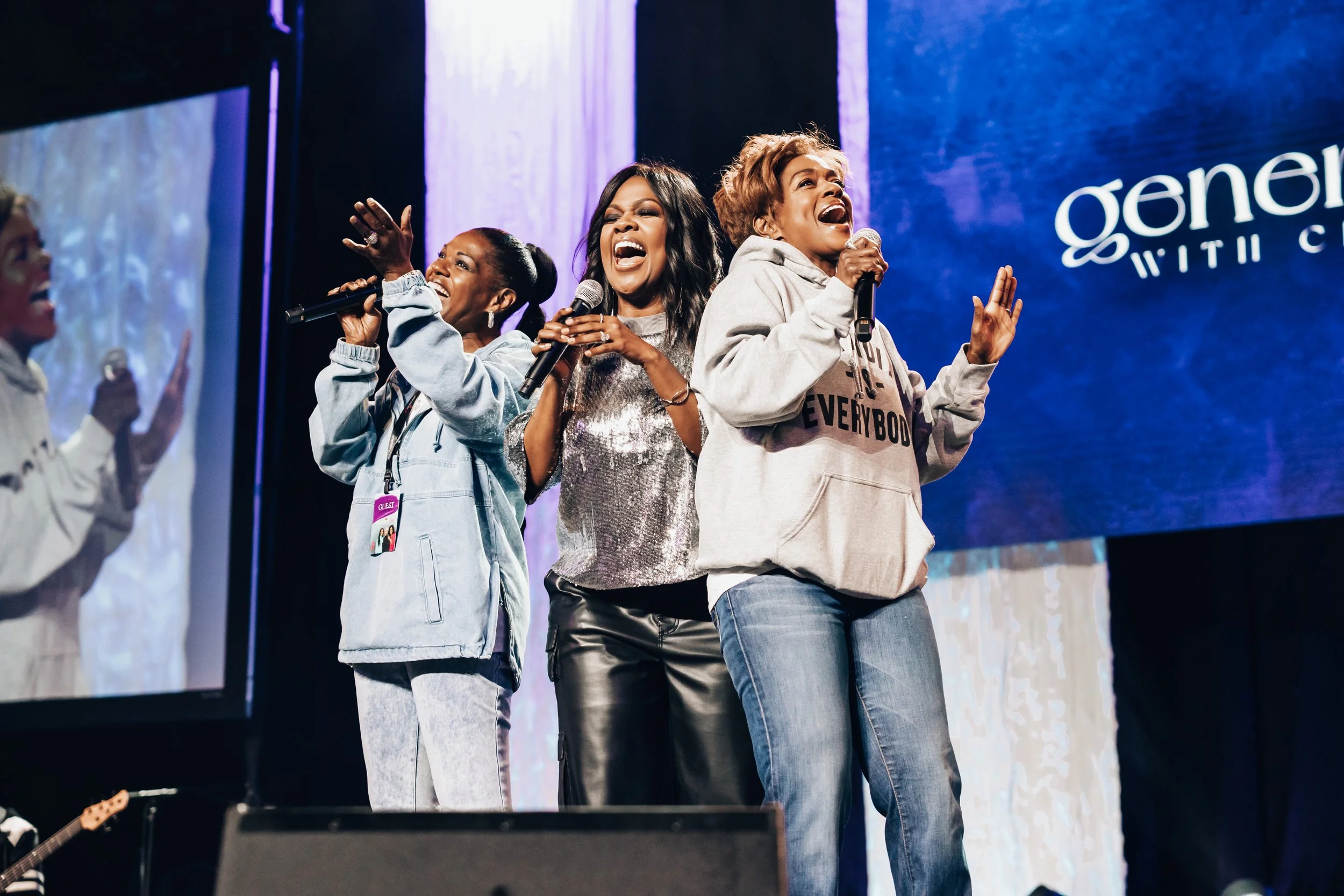 Three women singing and talking on stage at an event, with a large screen showing the logo 'generations with C...' in the background. One woman is wearing a shiny silver top, another is in a gray hoodie with text, and the third wears a denim jacket.