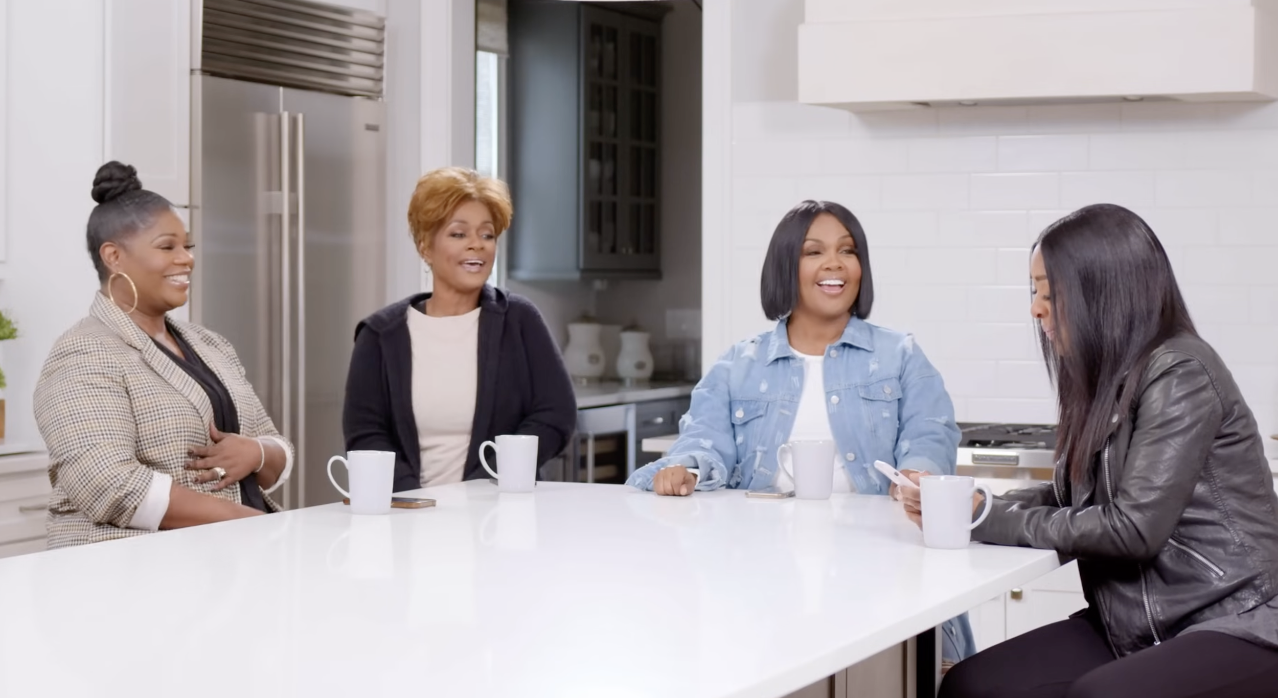 Four women sitting at a white kitchen island, smiling and talking, with three white coffee mugs on the table.