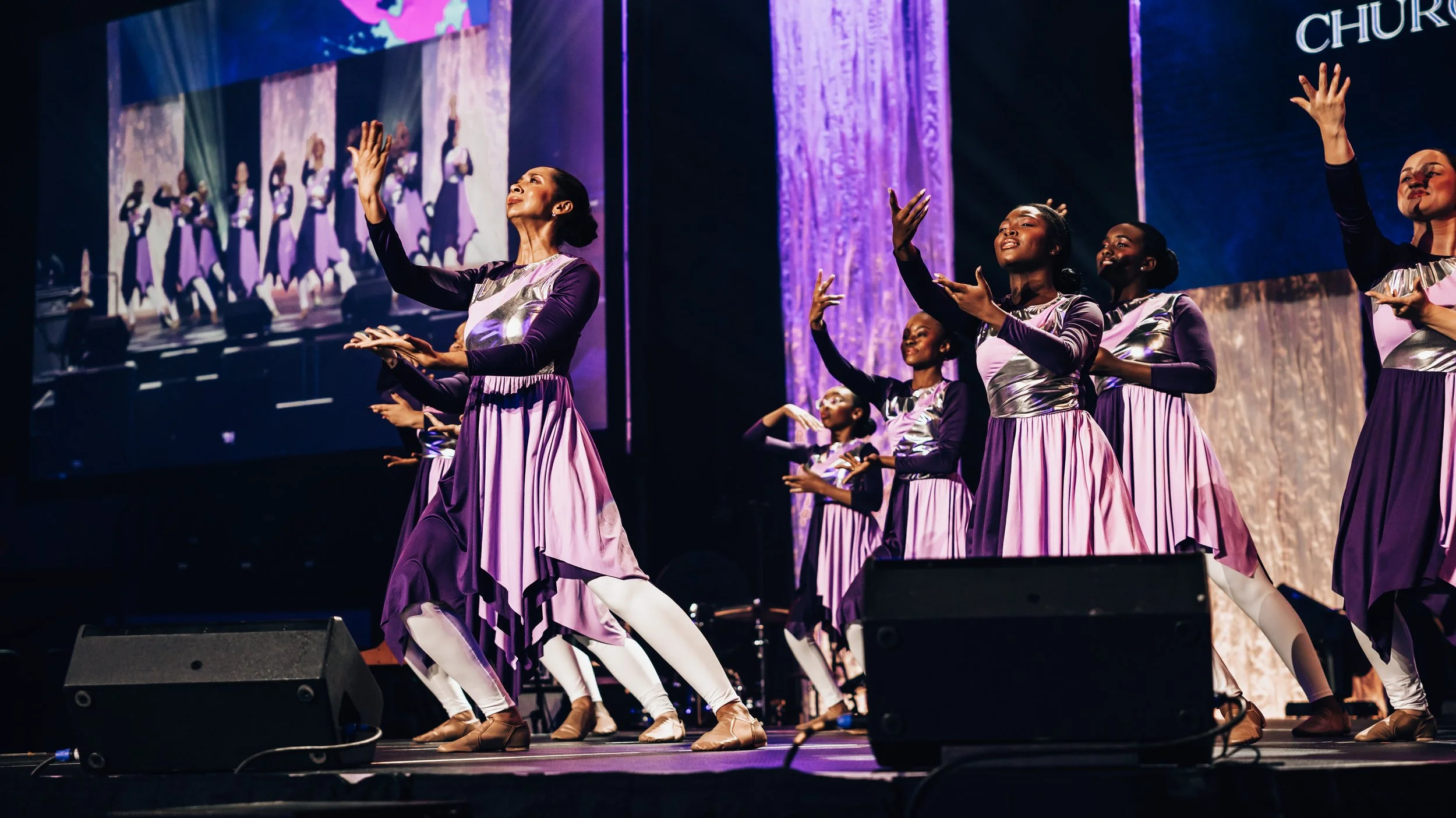 Group of women performing a dance on stage, wearing purple and silver dresses with white leggings, with a large screen showing their live performance in the background.