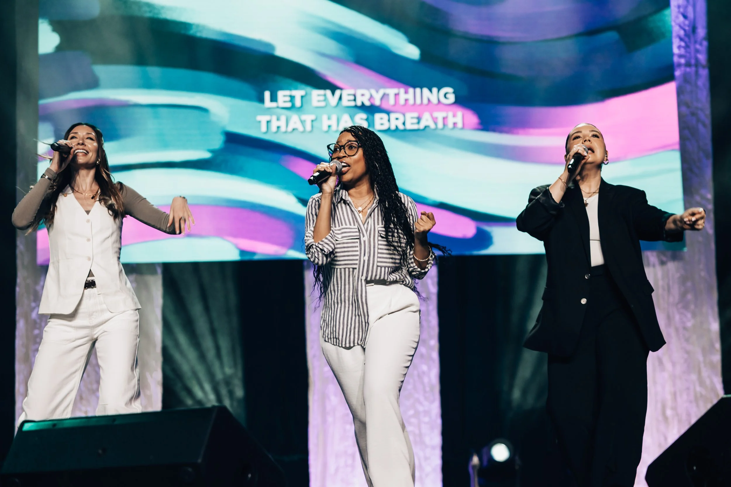 Three women singing on stage with a colorful background and a screen displaying the words "Let Everything That Has Breath".