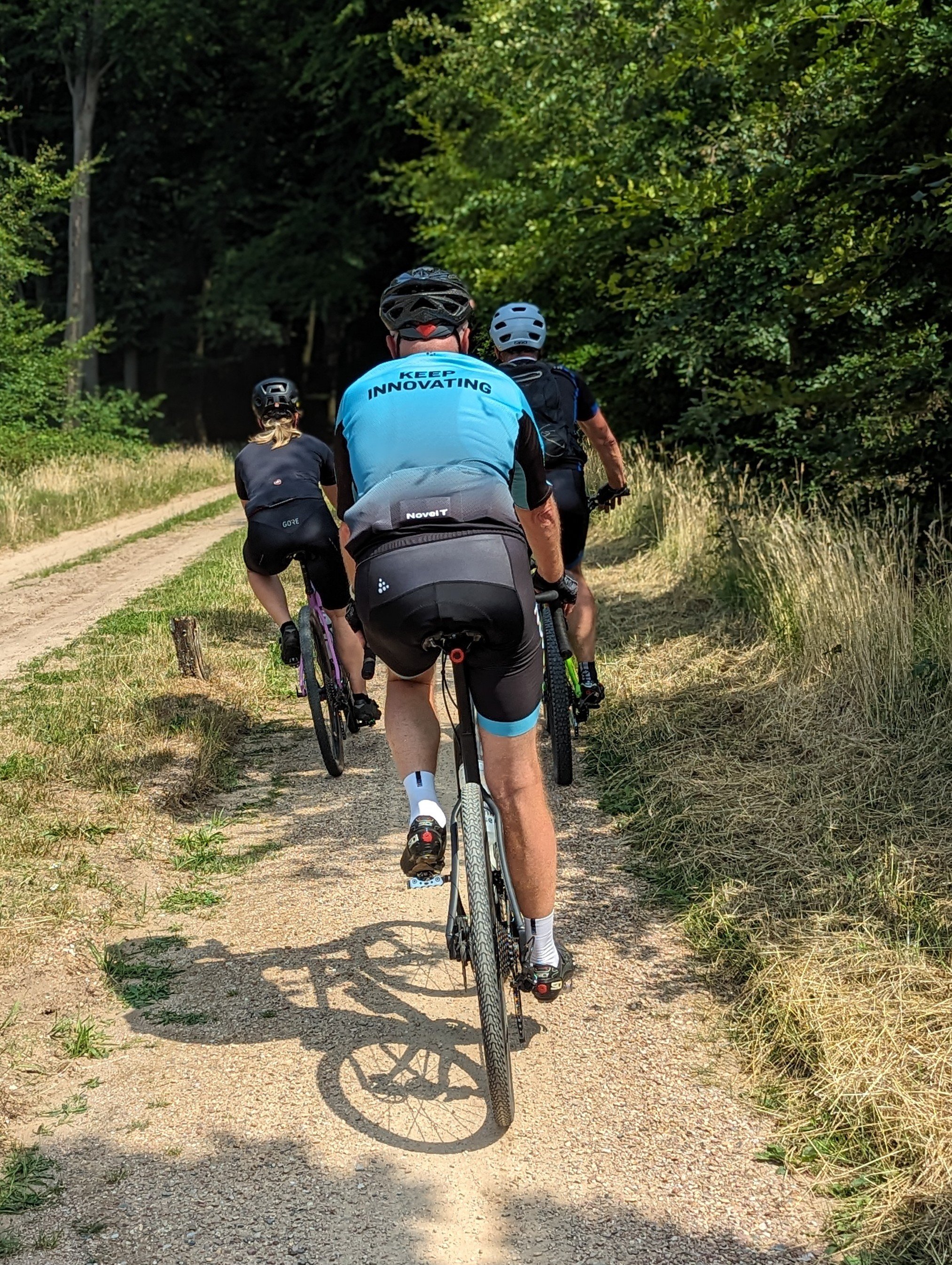 Drie mensen fietsen op een onverharde bosroute, omringd door groene bomen en gras.