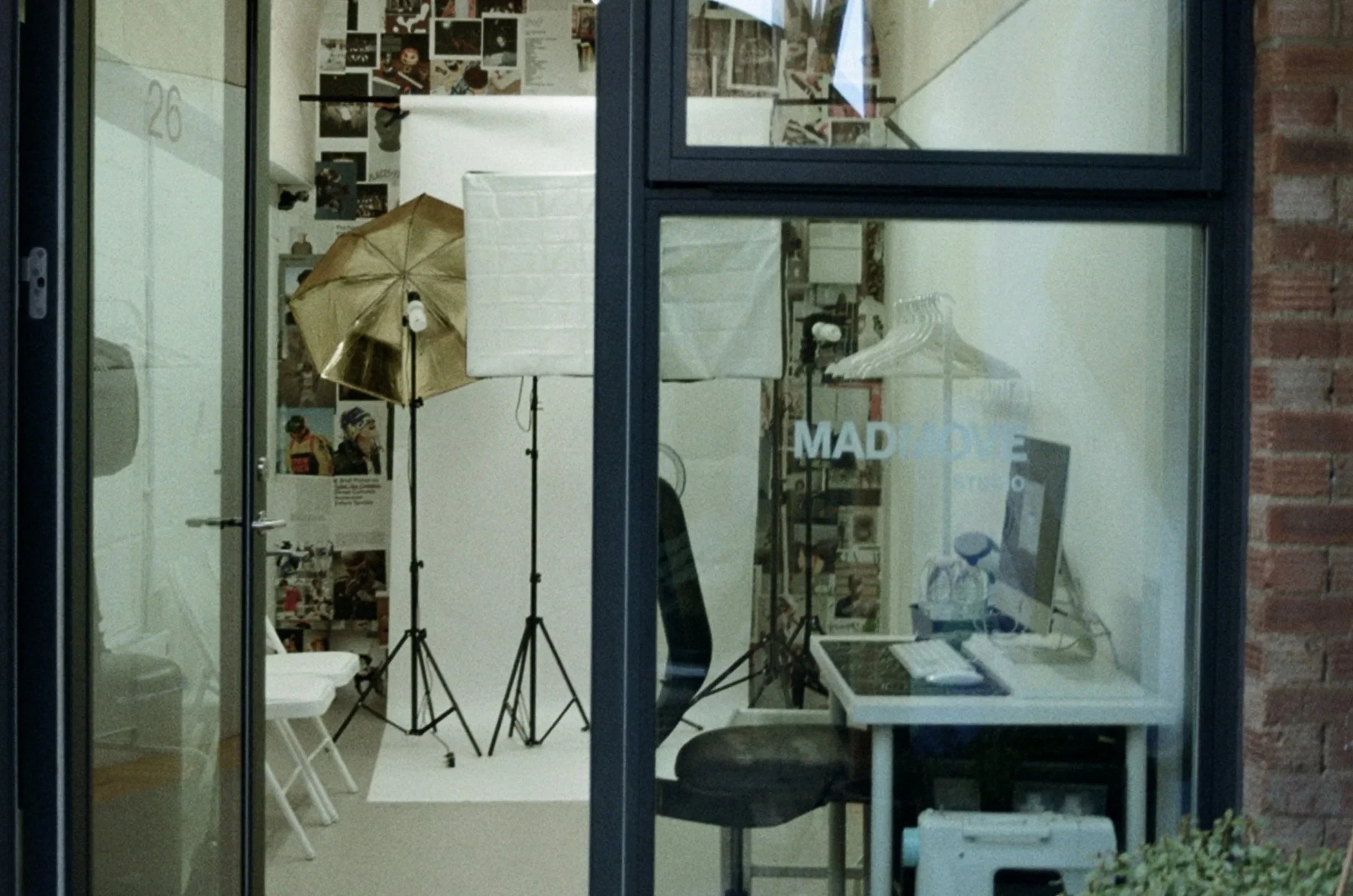View through a glass window of a photography studio with lighting equipment, a computer on a desk, and photographs on the wall.