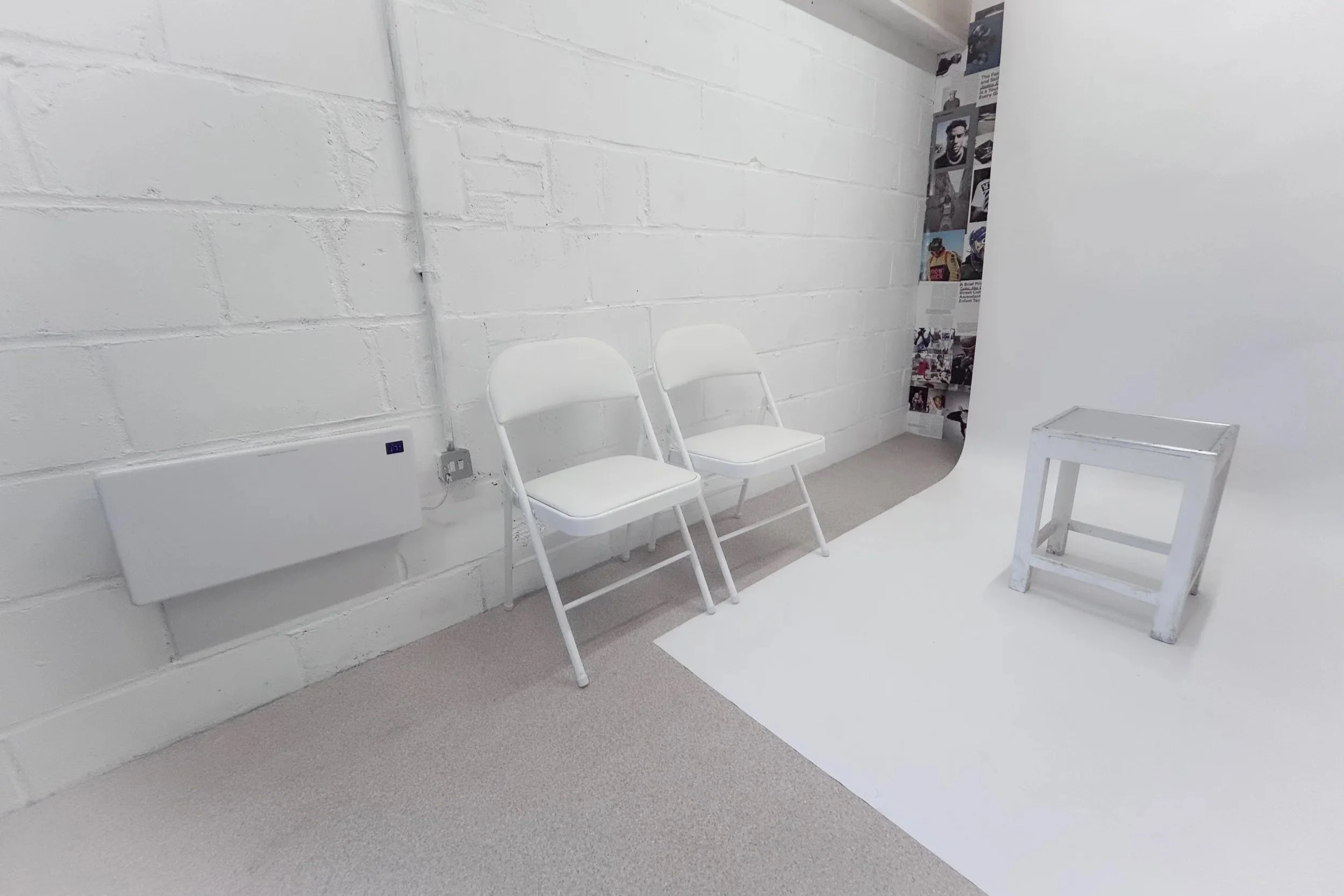 A photography studio with a white backdrop, two white folding chairs, a small white stool, and a collage of photos on the wall to the right.