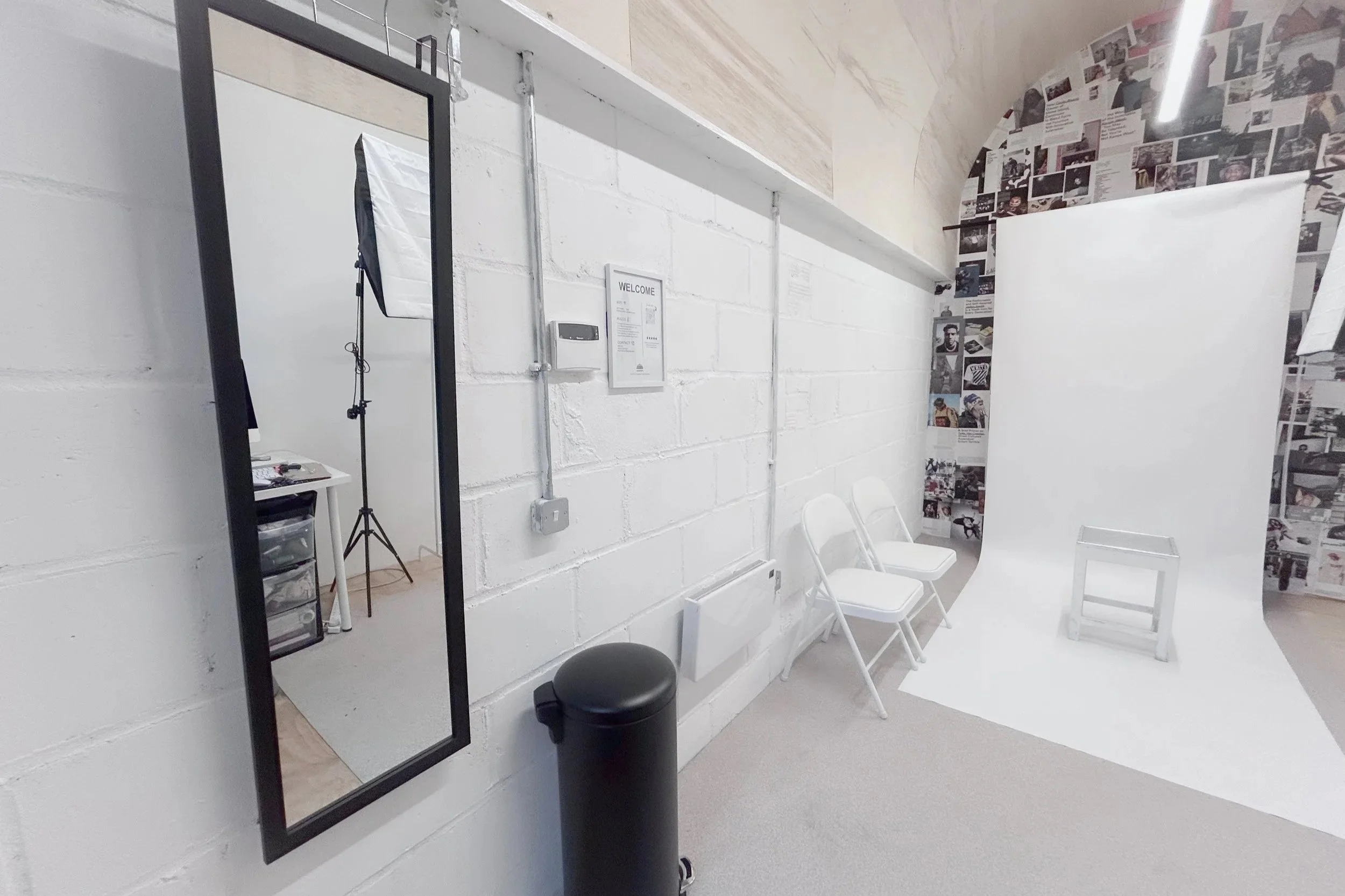 Photography studio with a white paper backdrop, three white chairs, a black trash can, a tall mirror, and a collage wall with photos.