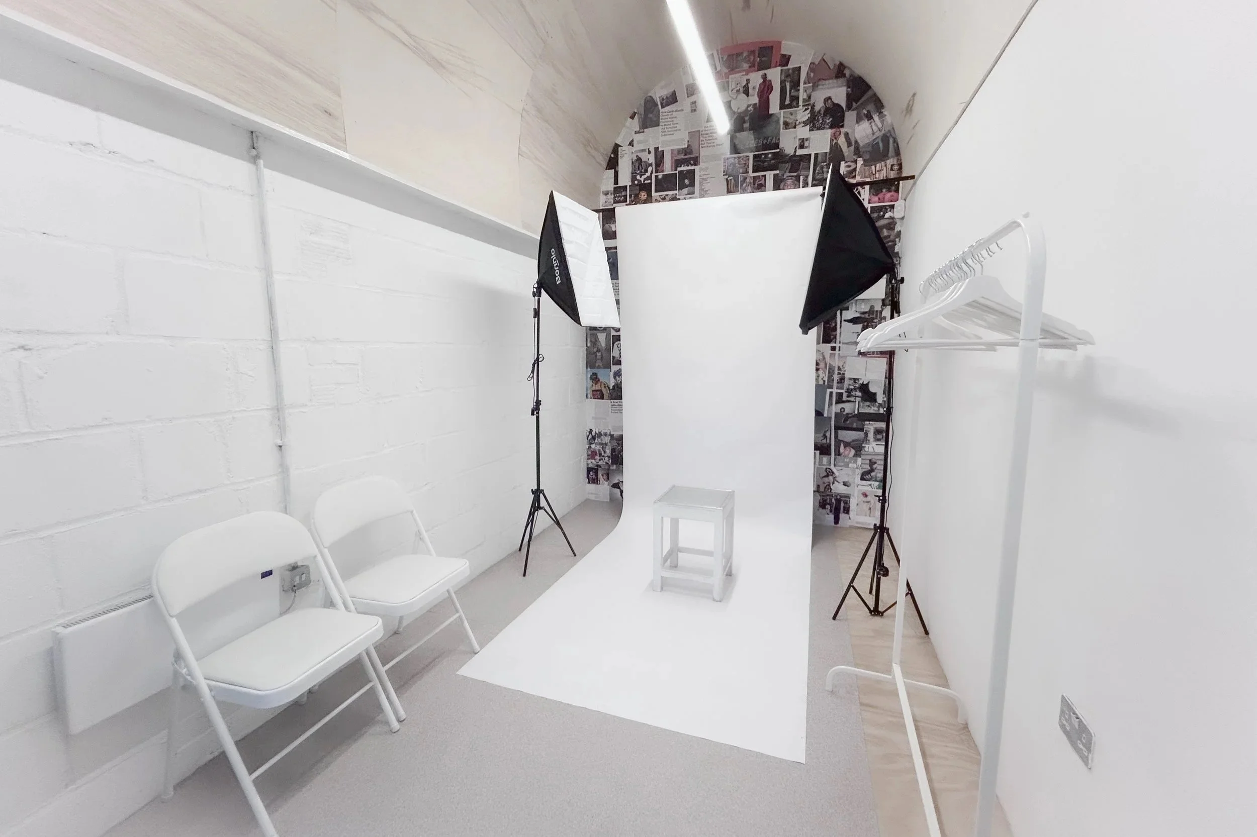 Photography studio with white backdrop, two softbox lights, a stool, two white chairs, and a white clothing rack in a room with white walls and a collage of photos on the back wall.