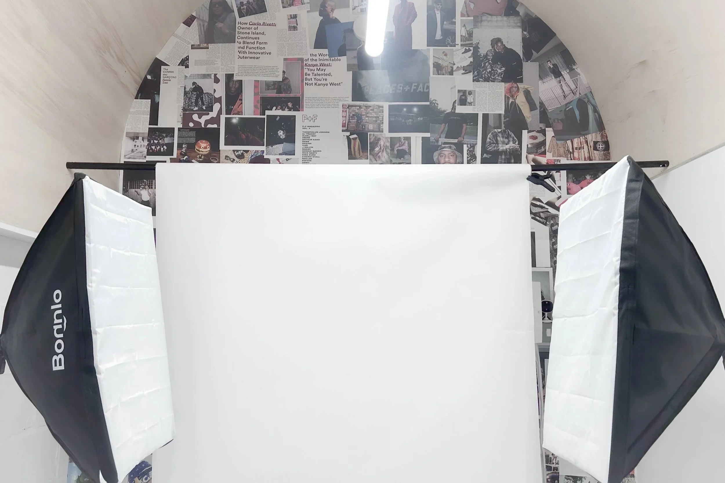 Photography studio setup with two softbox lights directed towards a white backdrop, and a wall covered with a collage of magazine clippings and photographs.