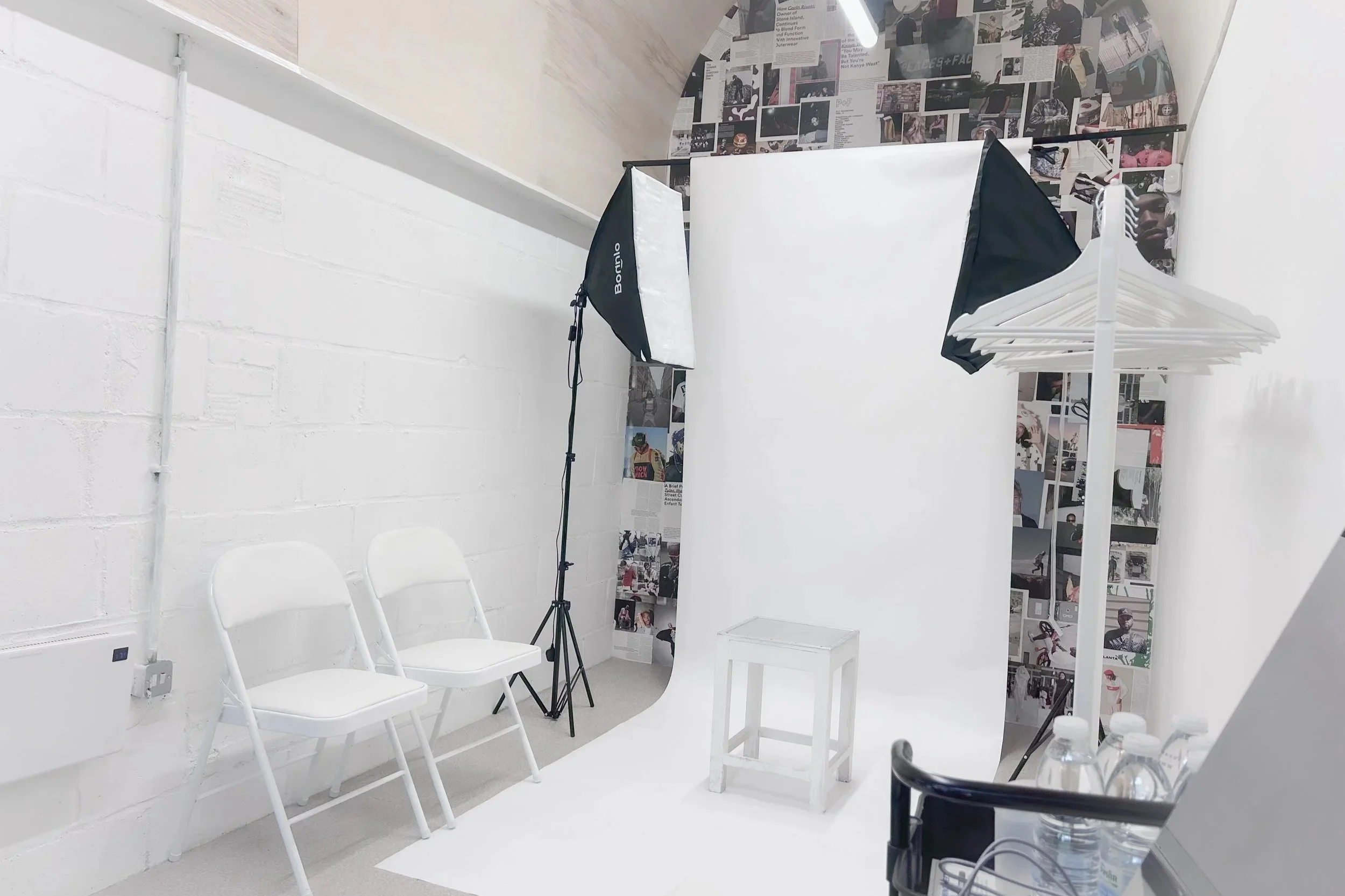 Photography studio with a white backdrop, studio lights, two white chairs, a small white stool, and a clothing rack with white hangers, decorated with collage posters on the wall.