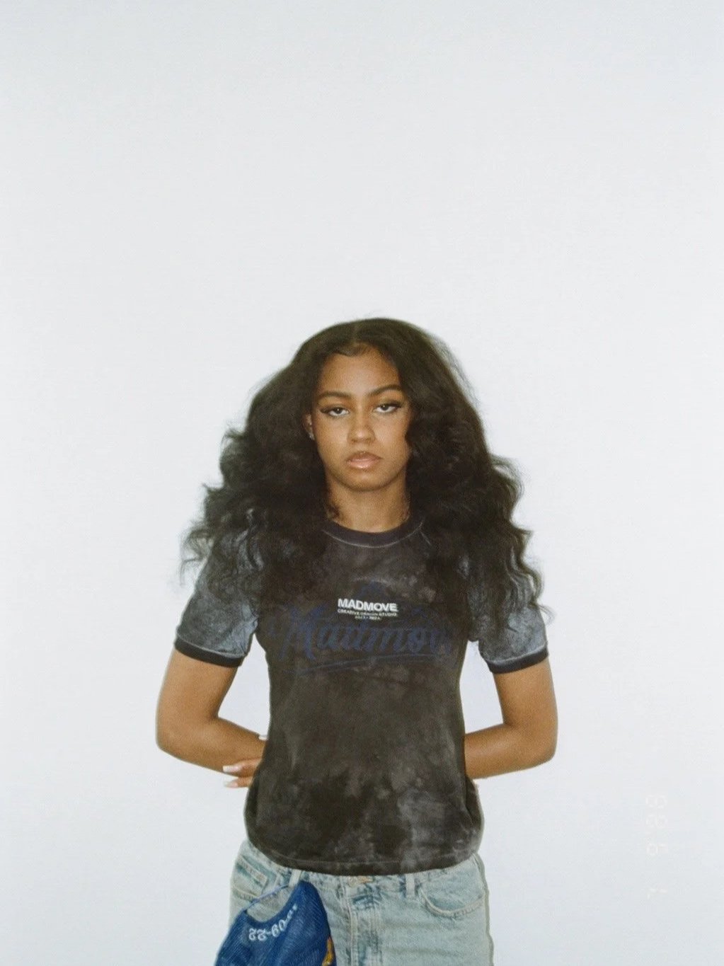 Young woman with long, curly hair standing against a white background, wearing a black tie-dye t-shirt and light blue jeans.