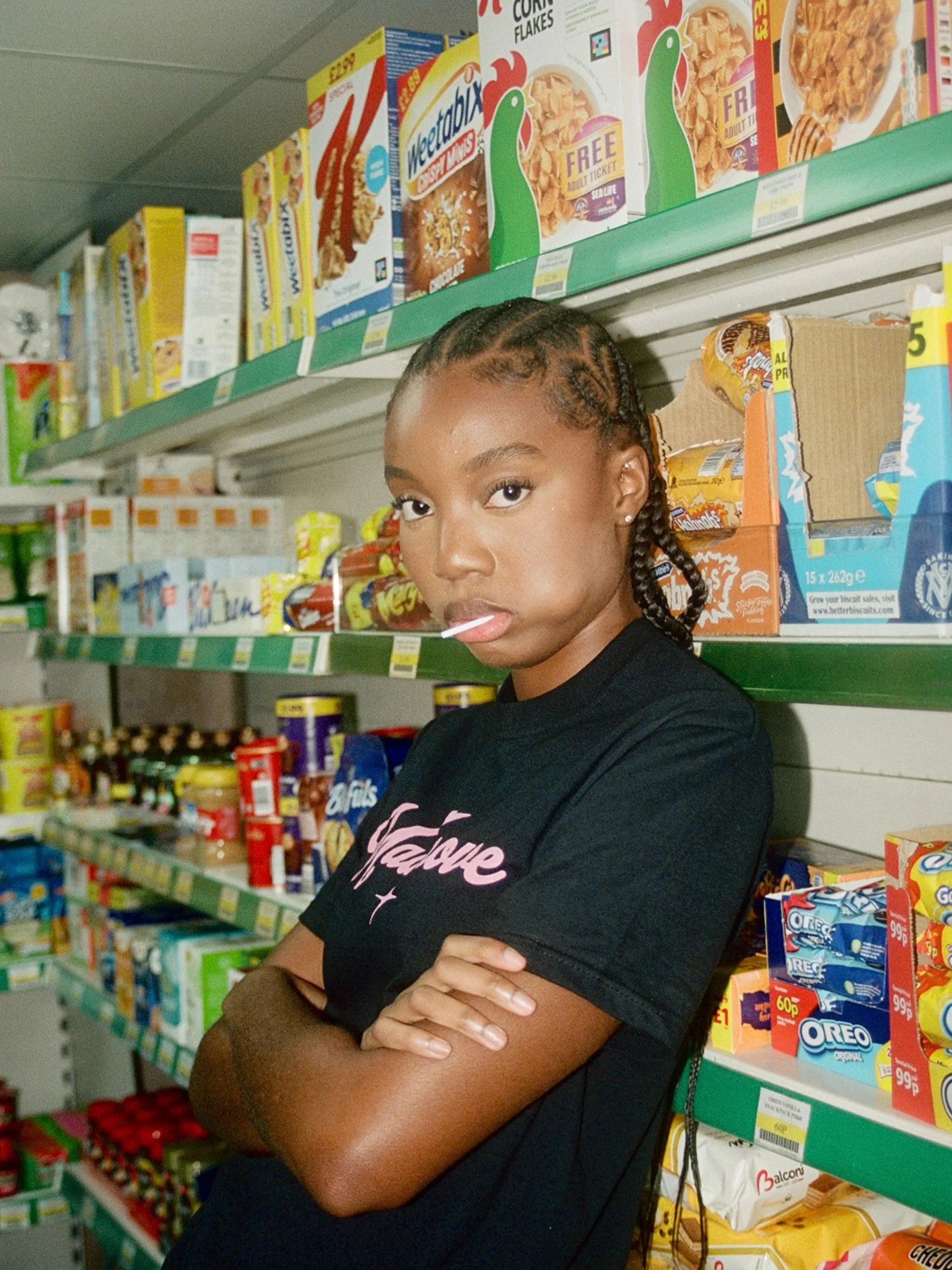 A young woman with braided hair stands in a grocery store aisle among shelves of cereal, with her arms crossed, looking into the camera.