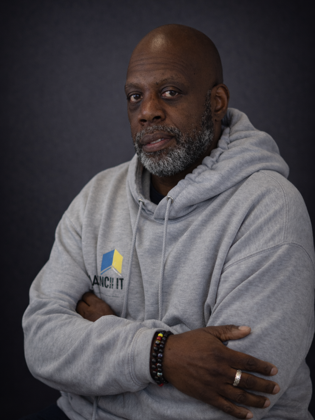 A middle-aged man with a gray beard and bald head wearing a gray hoodie with a logo on it, looking into the camera with arms crossed against a dark background.