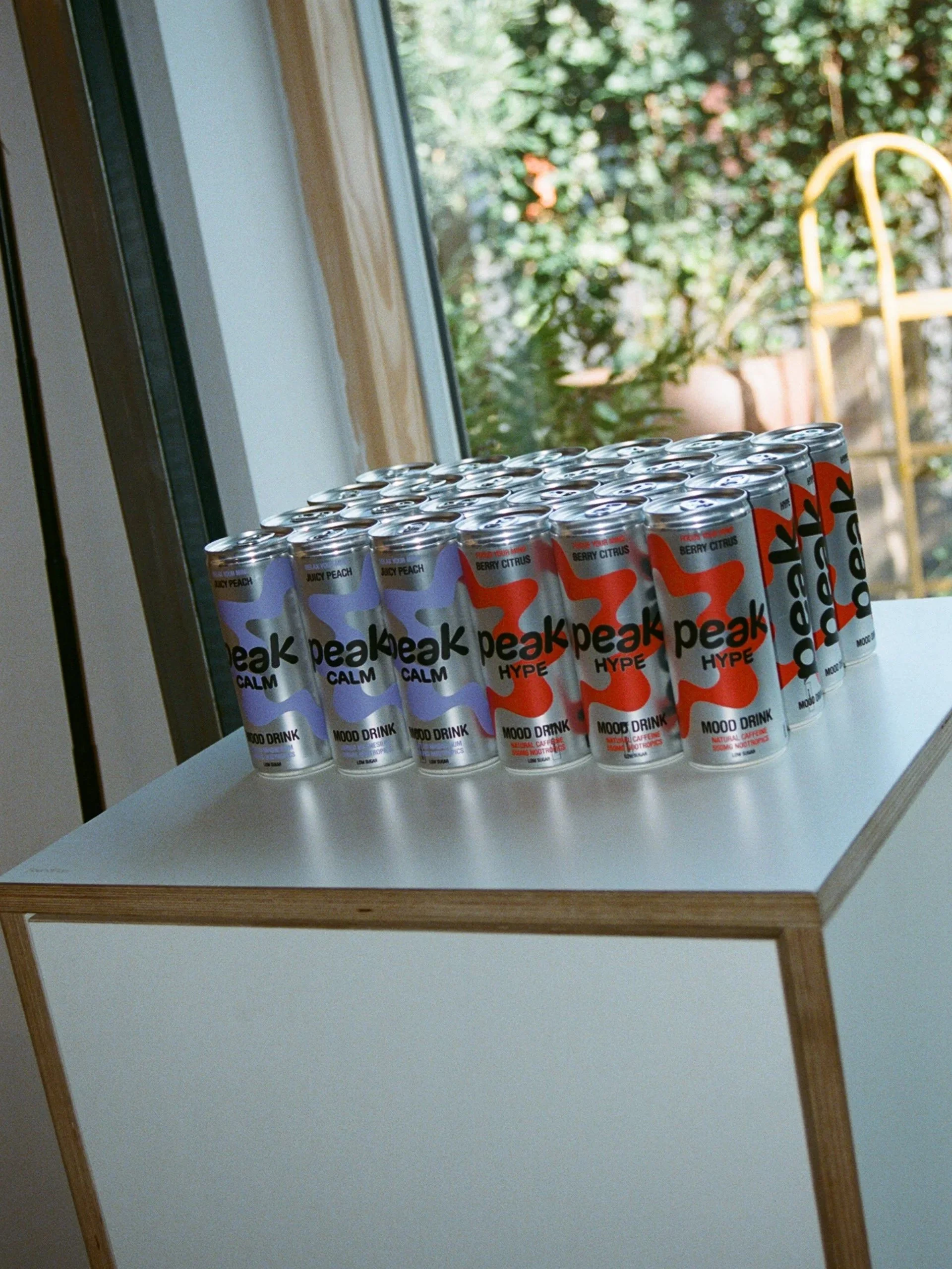 A row of canned sparkling water drinks with colorful labels on a white table, near a window with green foliage outside.