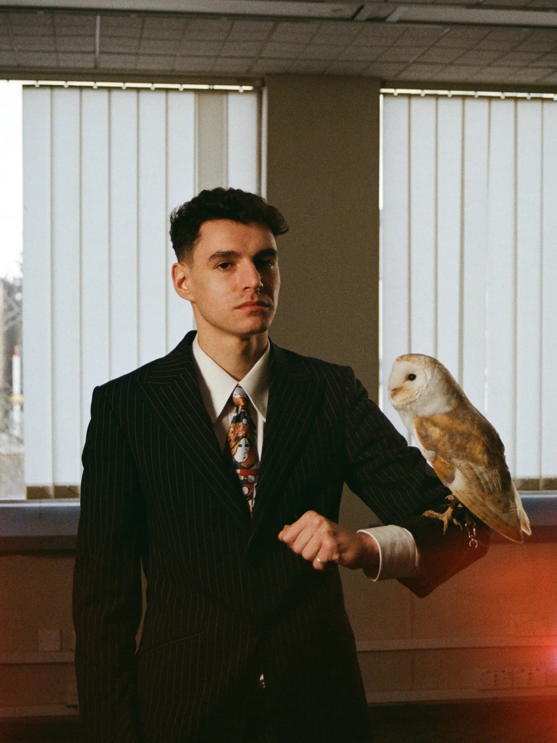 A young man in a dark pinstripe suit with a colorful tie, standing indoors with a barn owl perched on his arm.