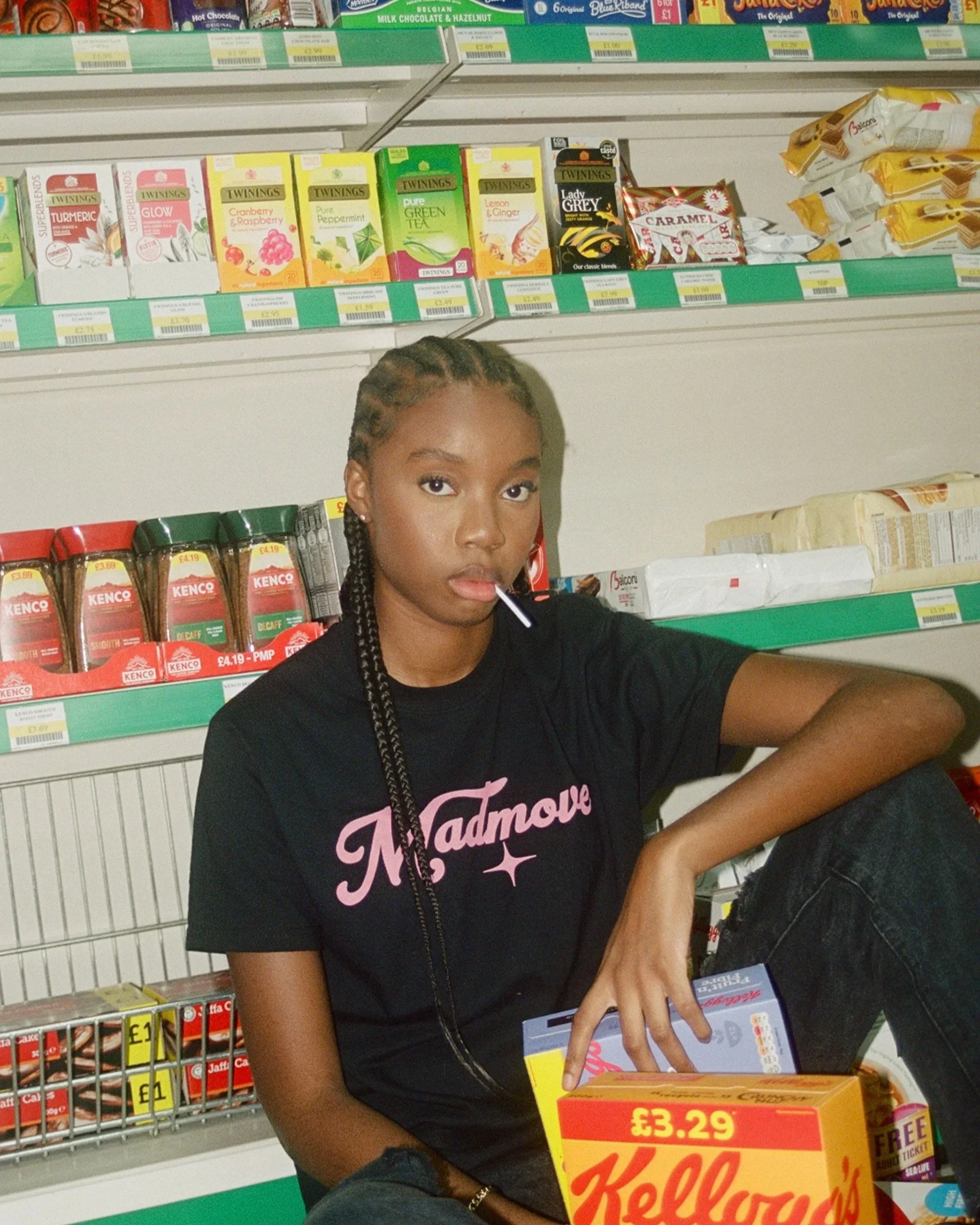 A young woman with braided hair sitting in a grocery store aisle surrounded by boxes of Kellogg's cereal and other food products. She is wearing a black T-shirt with pink writing and holding a box of Kellogg's cereal.