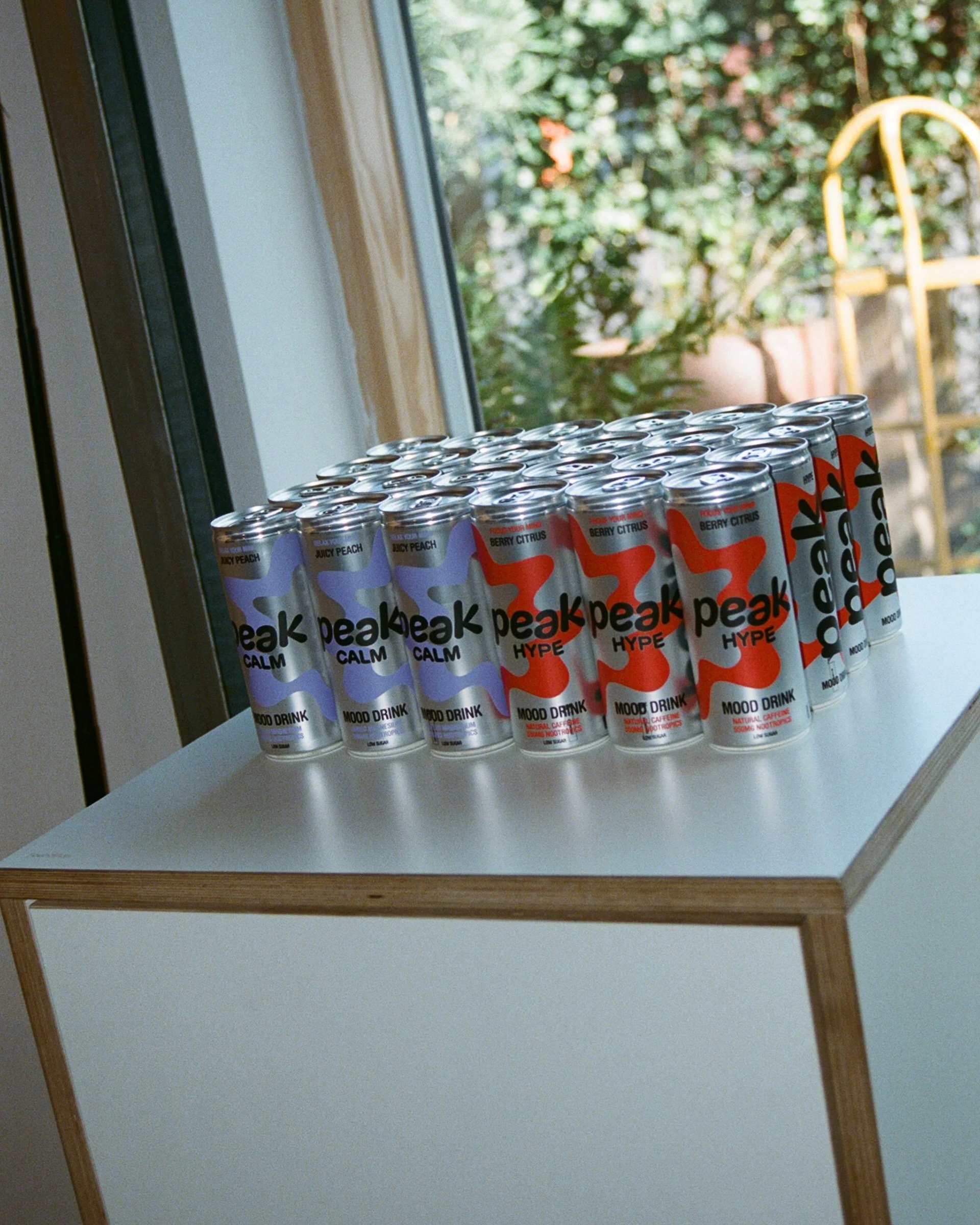 Row of cans of Peak mood drinks on a white table, with a window and outdoor greenery in the background.
