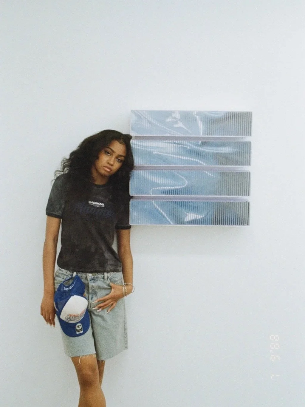 A young woman with dark, curly hair standing against a plain white wall, leaning slightly on a glass artwork. She is wearing a black graphic t-shirt and light denim shorts, holding a blue and white cap in her left hand.