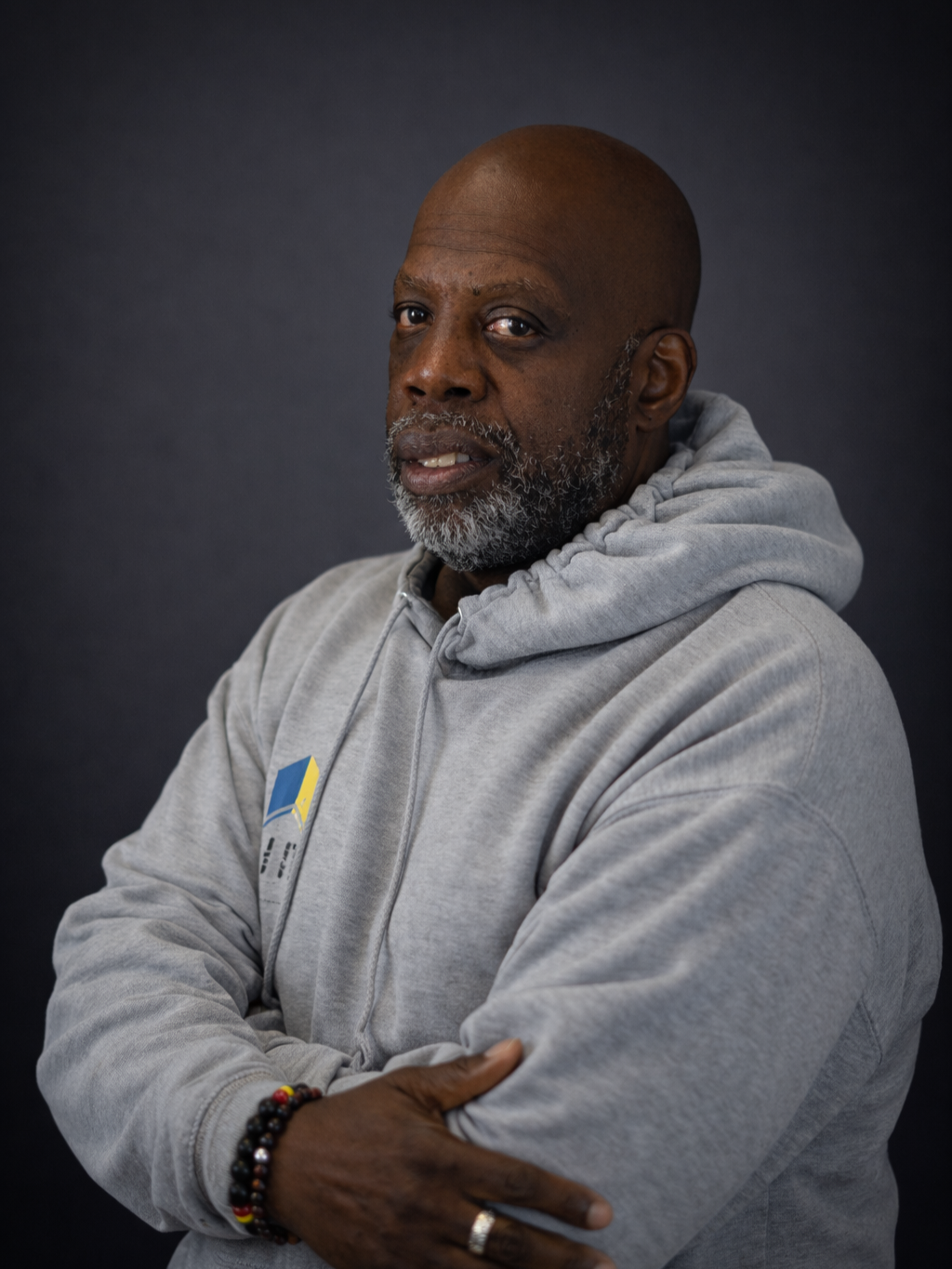 A middle-aged Black man with a gray beard wearing a gray hoodie, crossing his arms, against a black background.