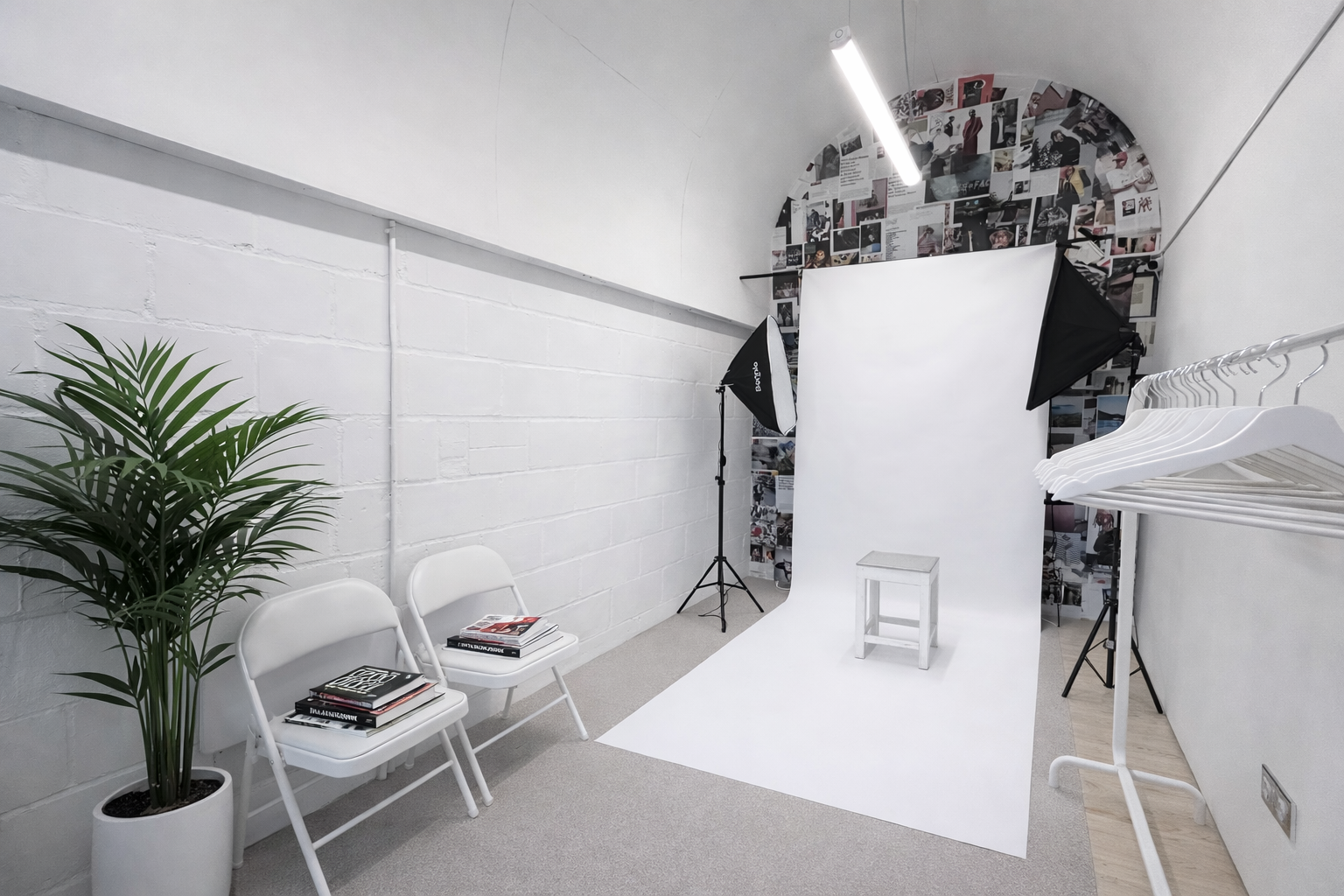 Photography studio setup with a white backdrop, studio lights, a small white stool, magazine stacks on white chairs, a potted plant, and hanging white clothes.