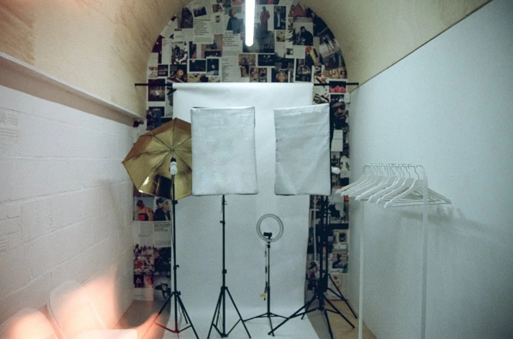 Photography studio setup with lighting equipment, diffusers, and a white backdrop in a room decorated with magazine pages on the back wall and white hangers on the right side.