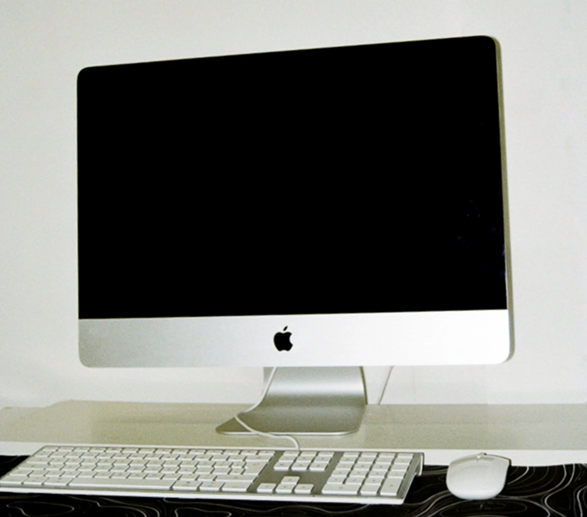 An Apple iMac computer with a matching wireless keyboard and mouse on a black desk mat against a white wall.