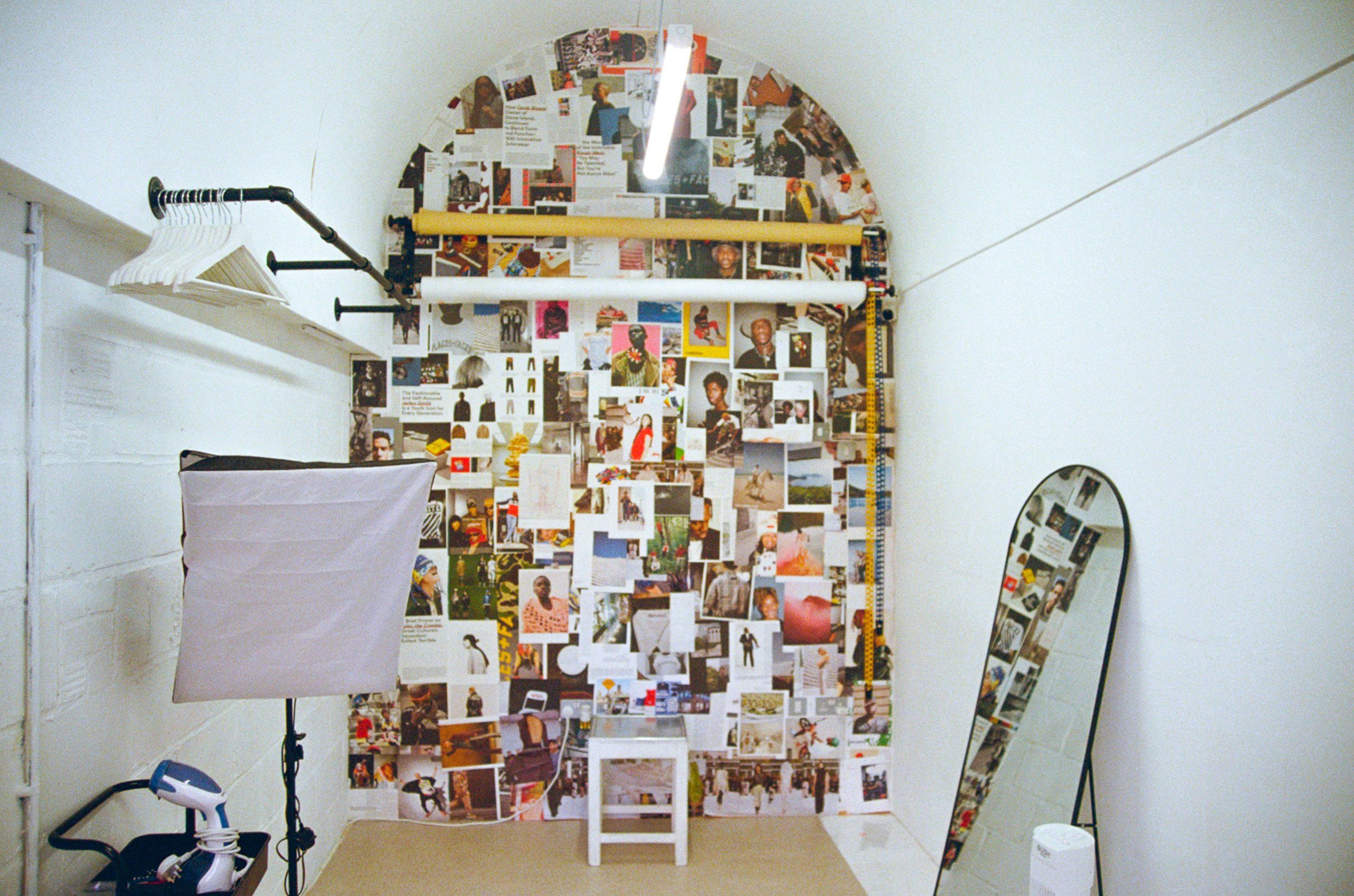 A collage of various photographs and images covers the back wall of an art or photo studio. To the left, there are some hangers on a rod, and below them, a lightbox. To the right, there is a large, tilted oval mirror with additional images reflected 