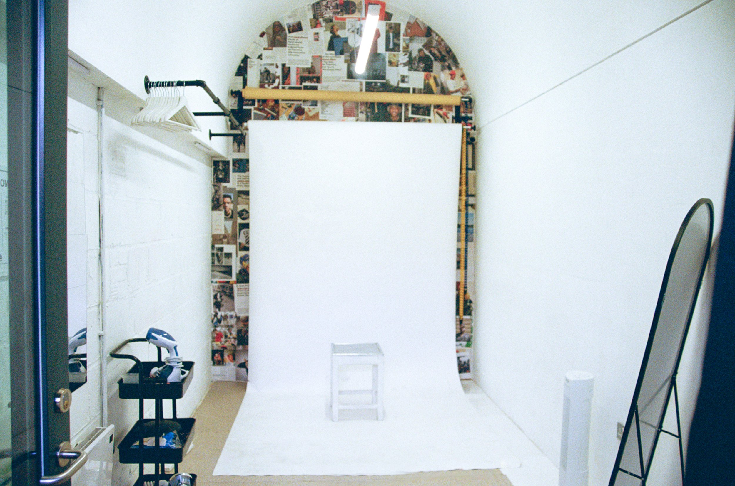 A photography setup with white backdrop, collage of magazine clippings on the wall, and various photography equipment including an iron, a black cart, and a stool inside a studio space.