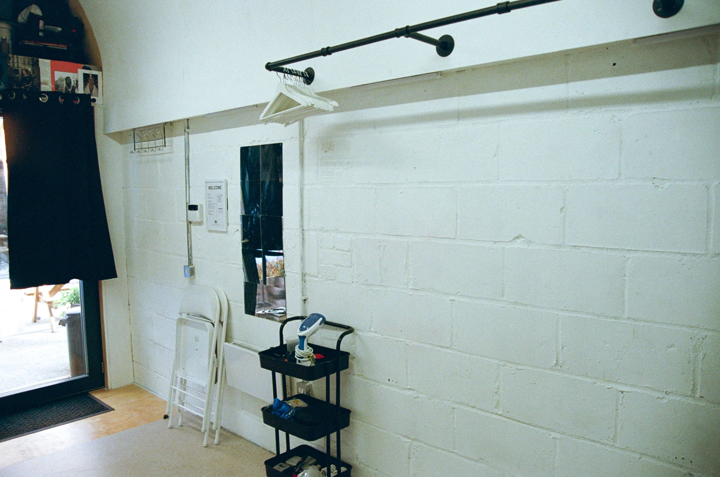 Interior of a room with white painted cinder block walls, showing a small black shelf unit with cleaning supplies below, a white ladder leaning against the wall, and a chair. Items are hung on a black rod attached to the wall, including white hangers