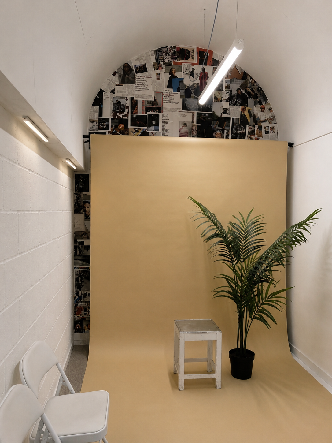 Photography studio setup with a beige backdrop, a potted palm plant, a small white stool, and white chairs, with a collage of black-and-white and color photographs on the wall above the backdrop.