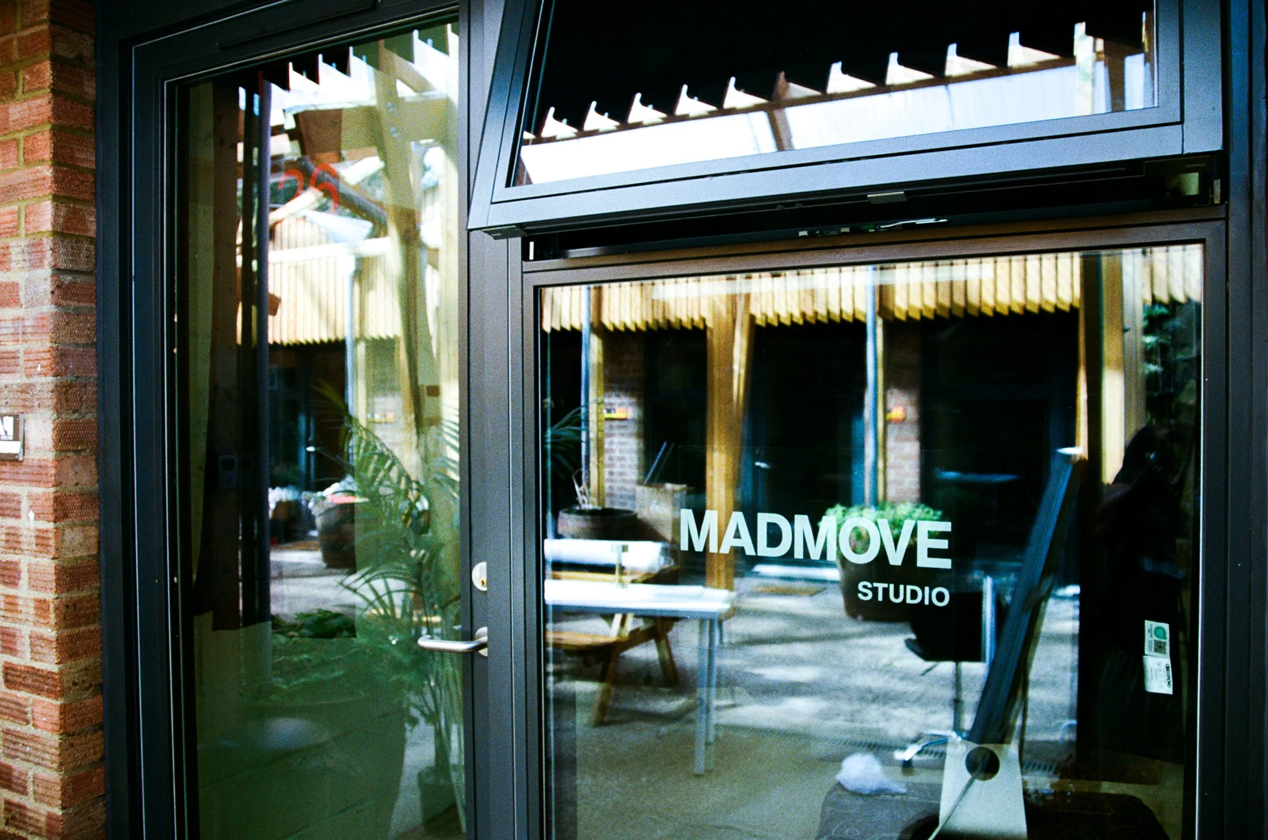 Glass door with the text 'MADMOVE STUDIO' visible, showing an interior space with tables and chairs, reflecting some trees and outdoor elements.
