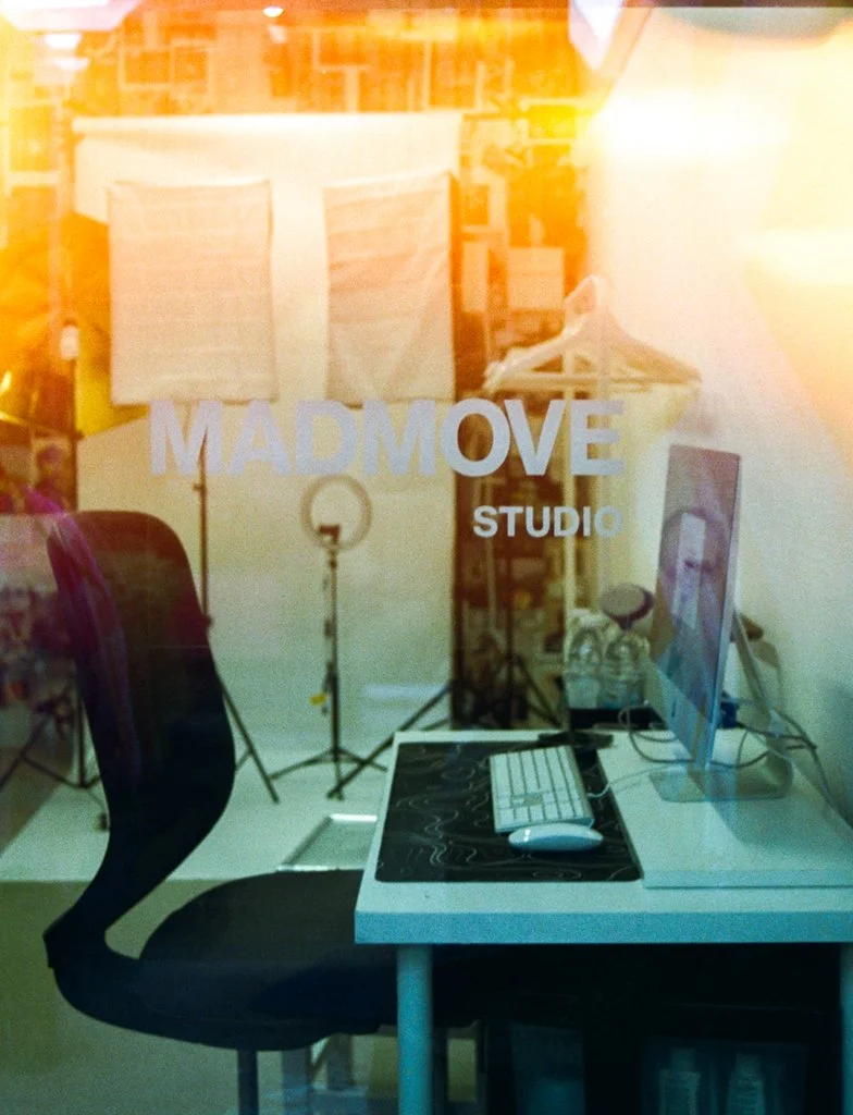 Photo of a creative workspace viewed through a glass door with lettering that reads 'Mad Move Studio,' showing a computer on a desk, a chair, and photography or filming equipment in the background.