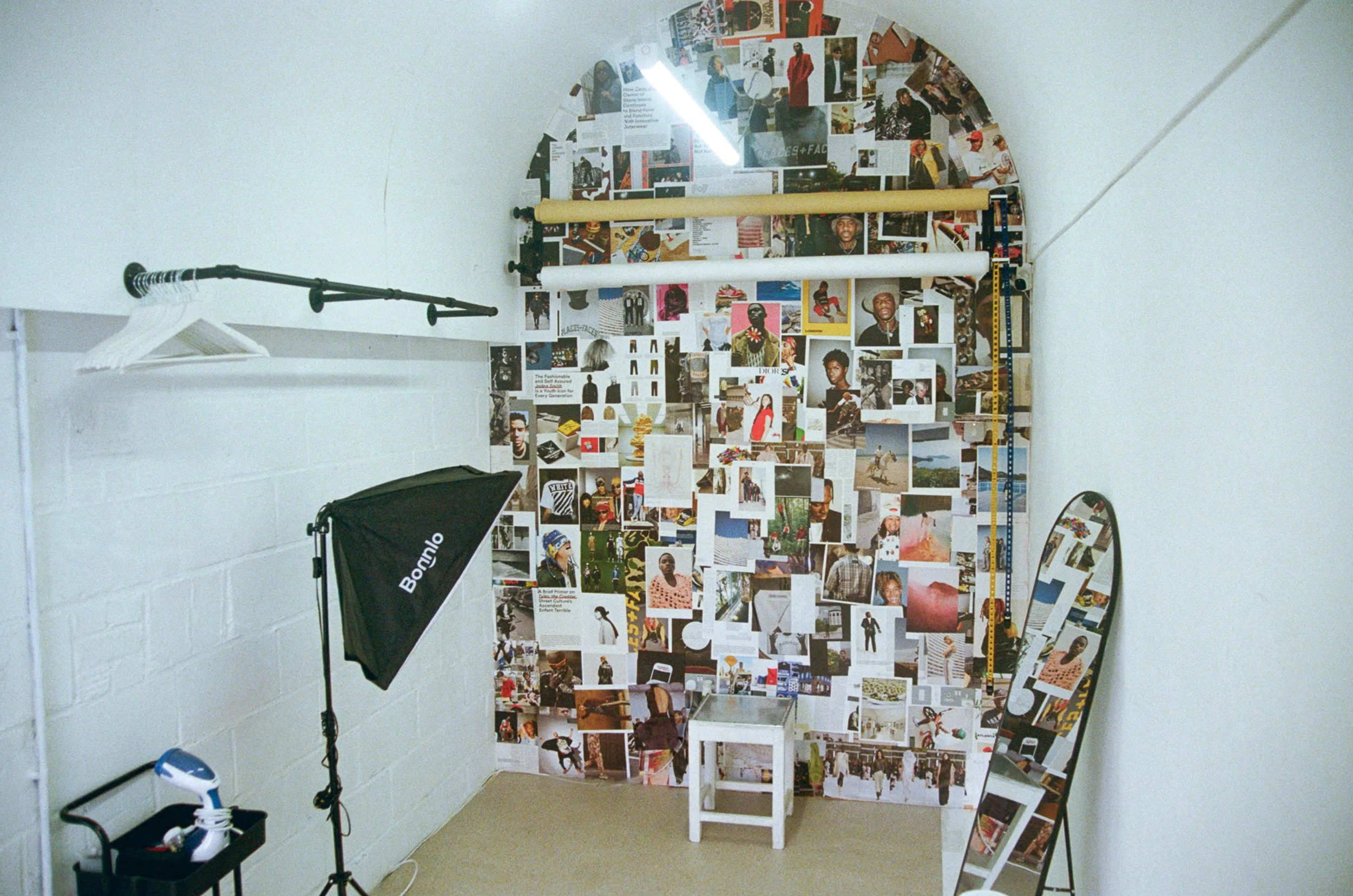 A wall covered with a collage of photographs and magazine cutouts of various people, along with some text. There are lighting and photography equipment, including a softbox and a reflector, in the room.