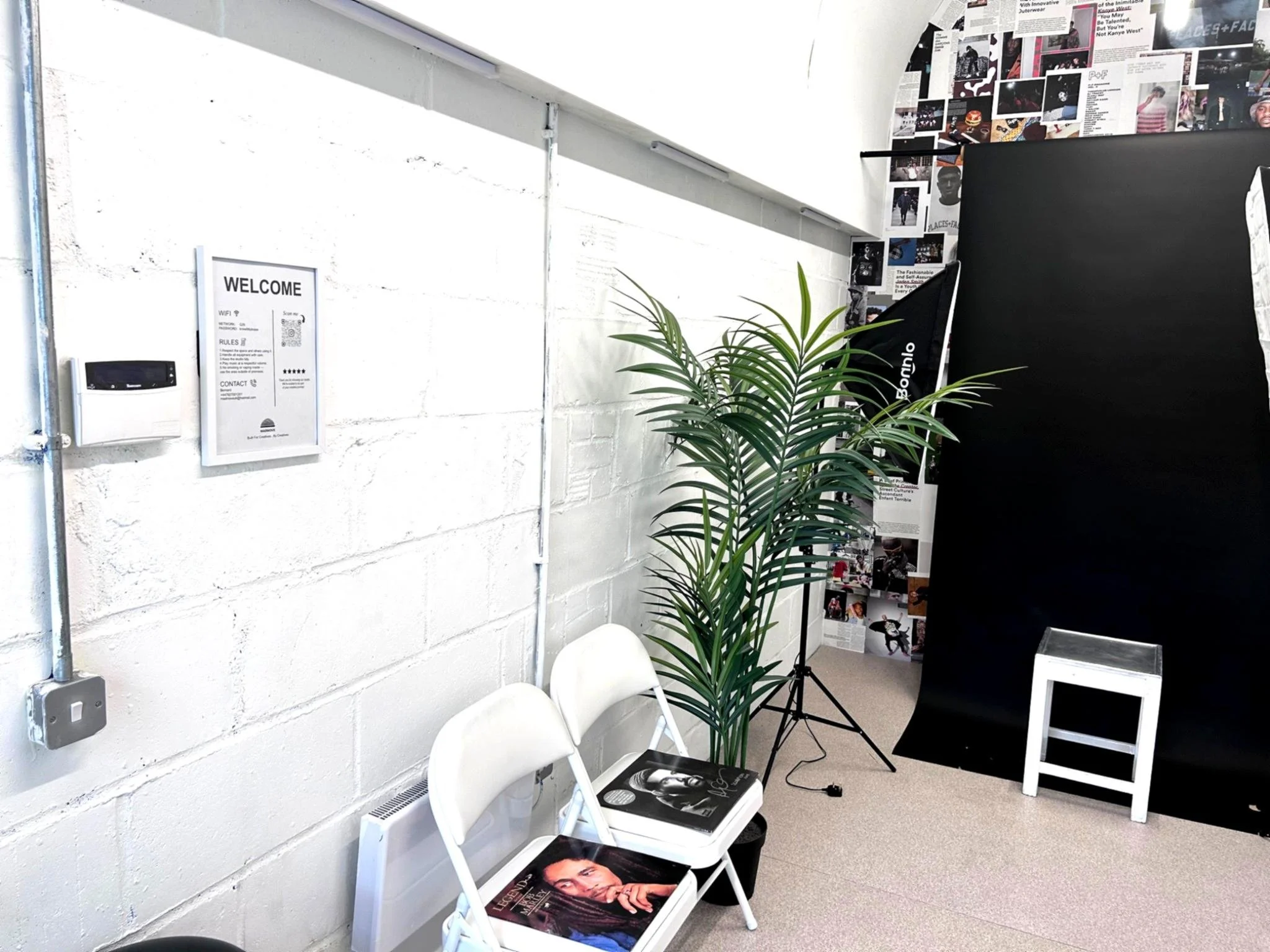 Gallery corner with white chairs, a tall plant, a black display board, and a collage of magazine pictures on the wall.