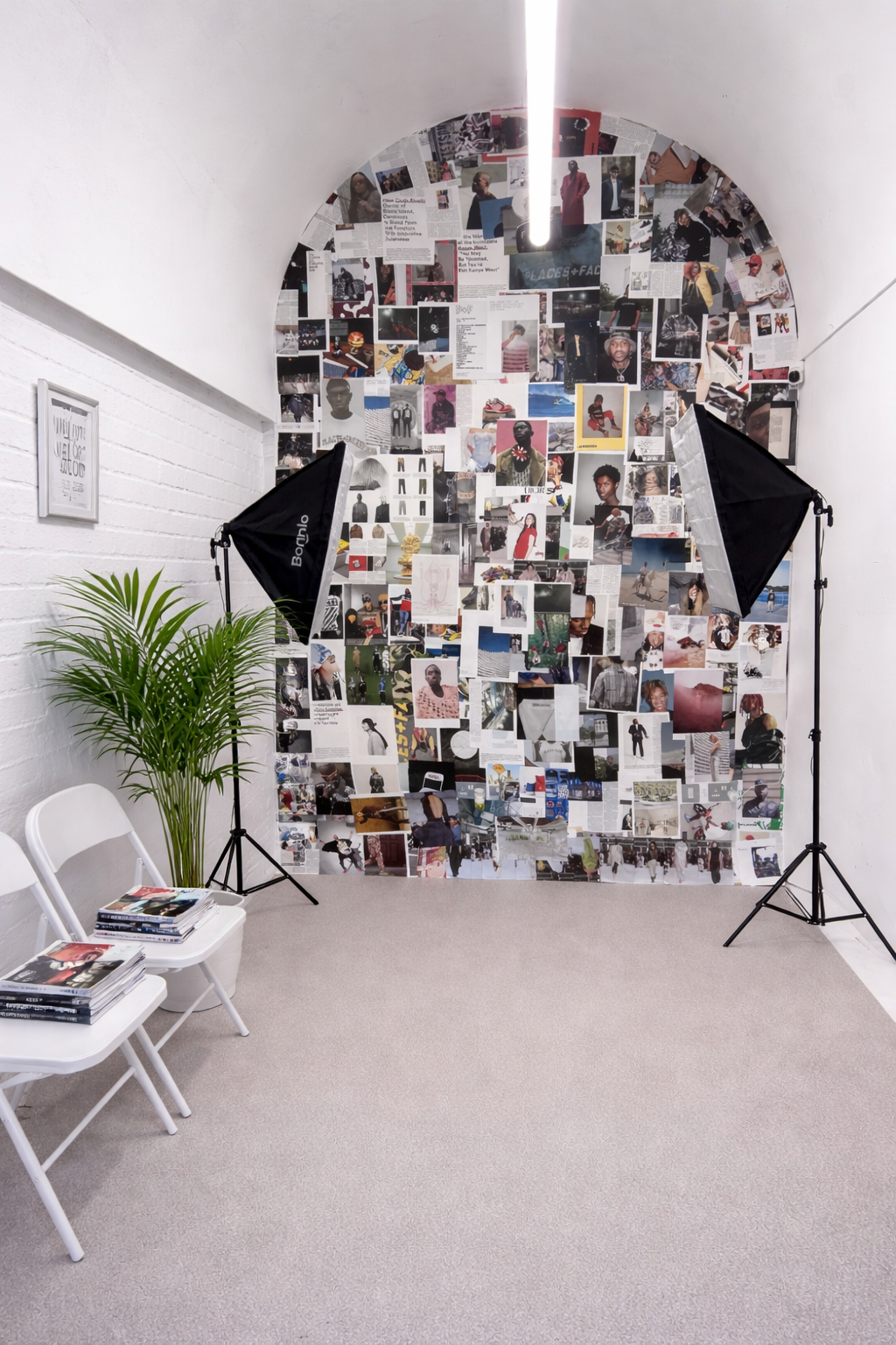 Photo studio setup with a collage wall of fashion photos and articles, two black photography umbrellas, a potted plant, and two white chairs with magazines.