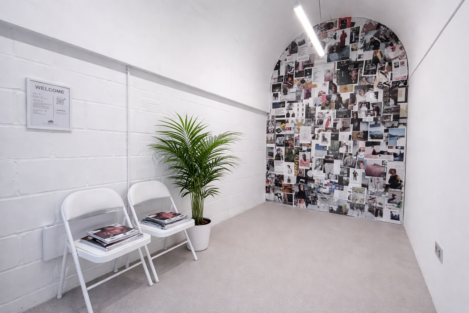 Waiting area with two white chairs stacked with magazines, a tall potted plant, a white brick wall with a welcome sign, and a collage of photos and paper on a curved wall.