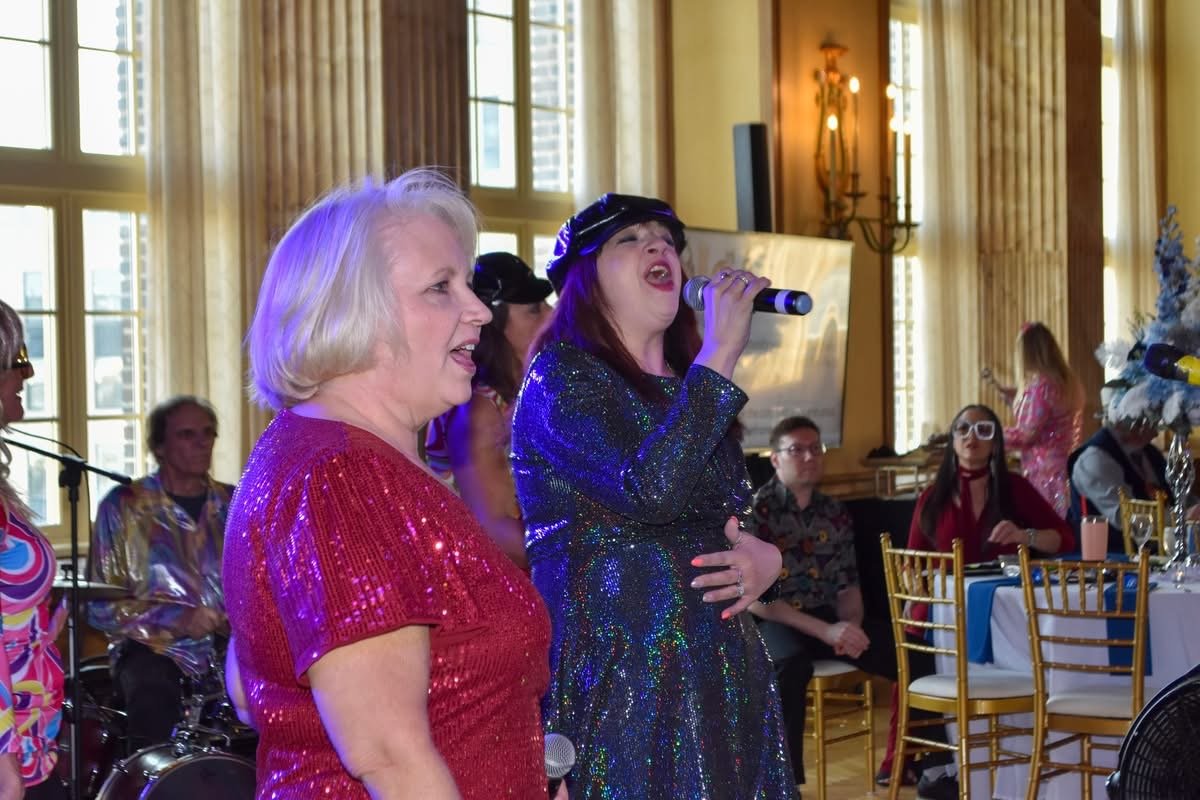 Two women singing at an indoor celebration, one wearing a red sequined top, the other in a glittery dark dress and cap, with musicians playing in the background and people seated at tables.