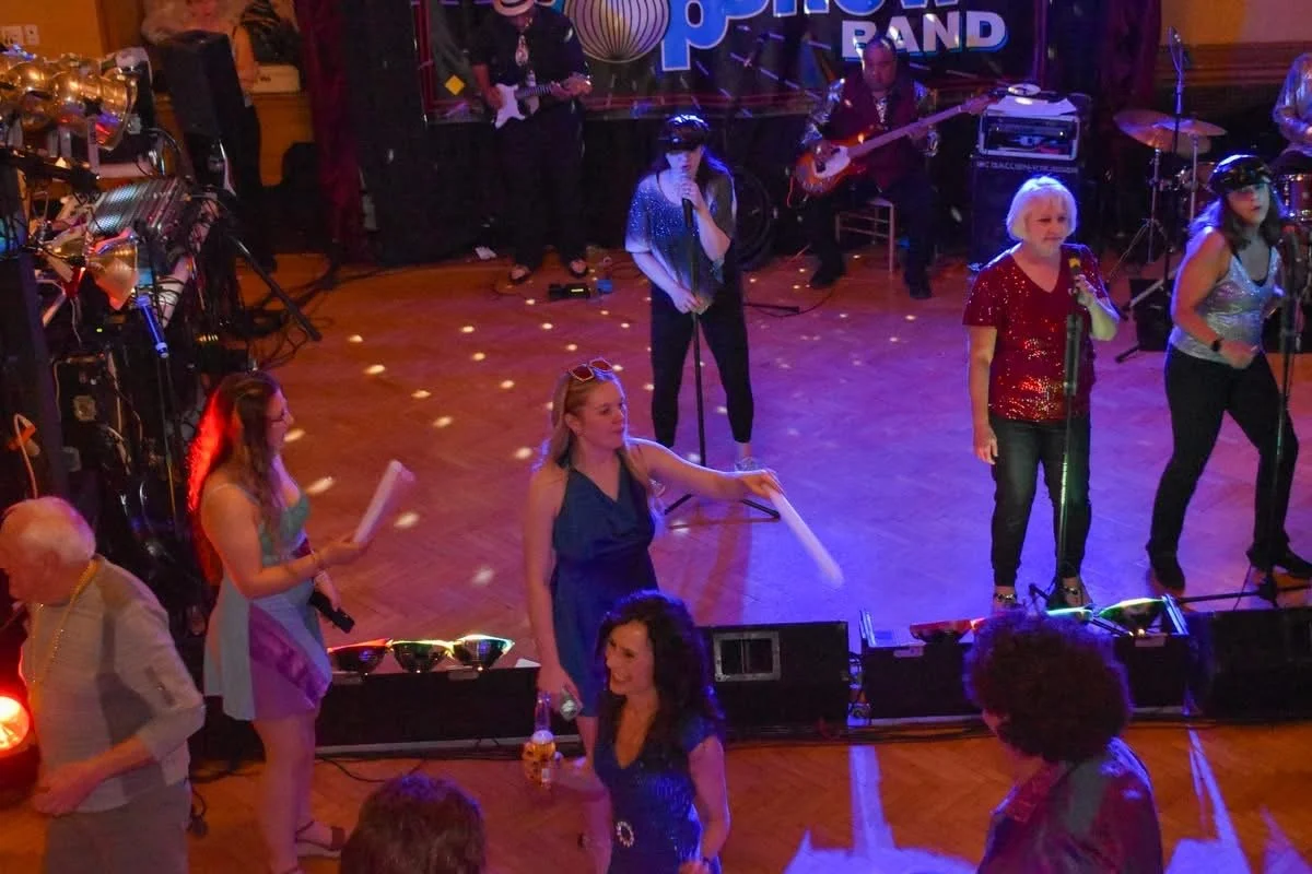 People performing on stage at a band event in a decorated indoor venue, with some audience members watching and dancing.