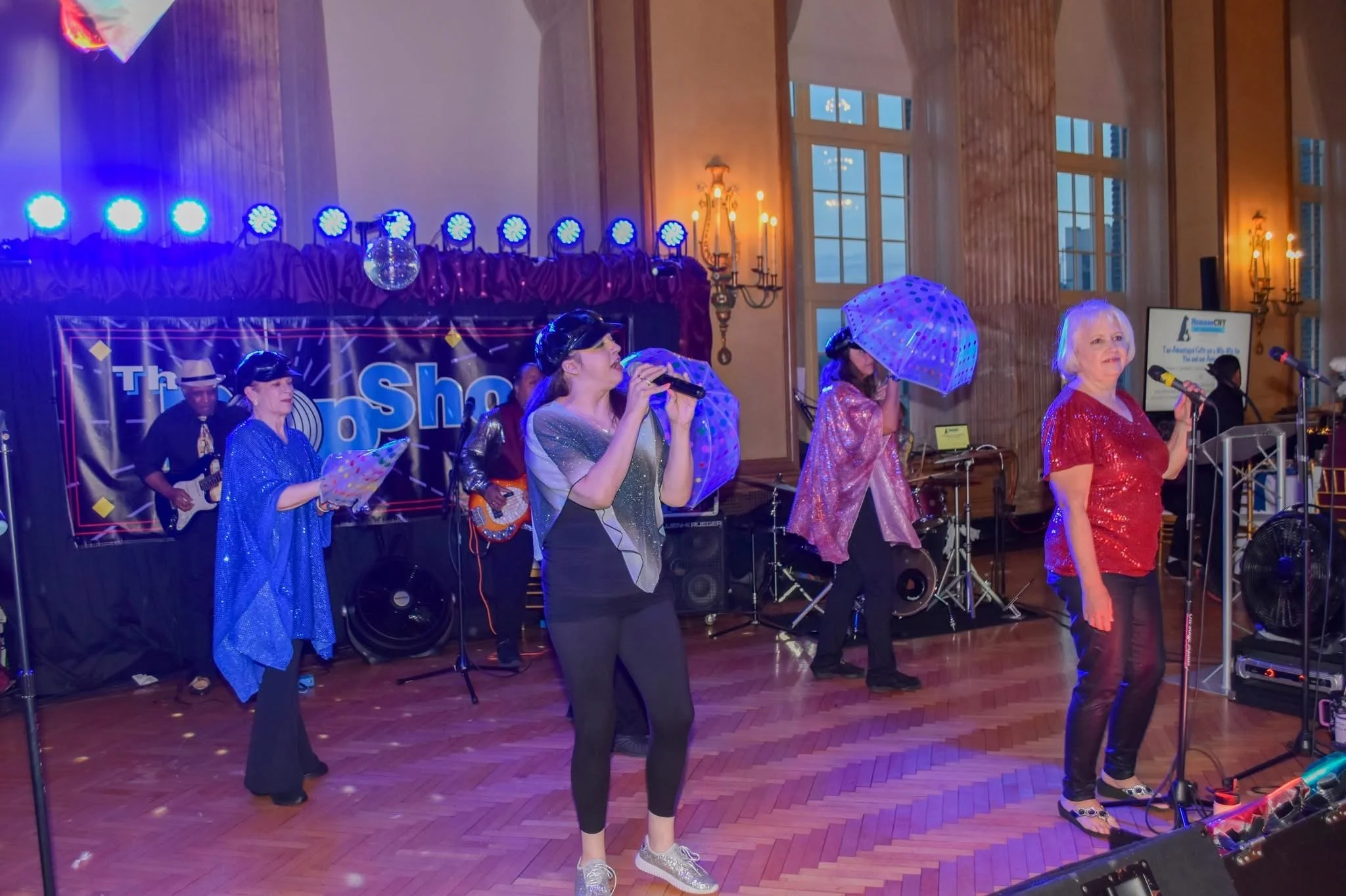 People performing on stage at an indoor event, with musical instruments, microphones, and colorful stage lighting, including women singing and playing instruments, and a man playing guitar in the background.