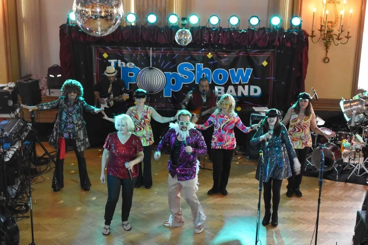 A band named 'The Pop Show Band' performing on stage with seven musicians dressed in colorful, 70s-style costumes and sunglasses, with musical equipment and stage lights.