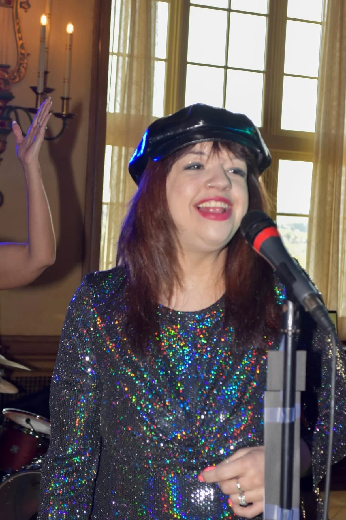 A woman with red hair singing into a microphone, wearing a shiny, multicolored, sequined top and a black leather cap, indoors with large windows and a candelabra on the wall behind her.