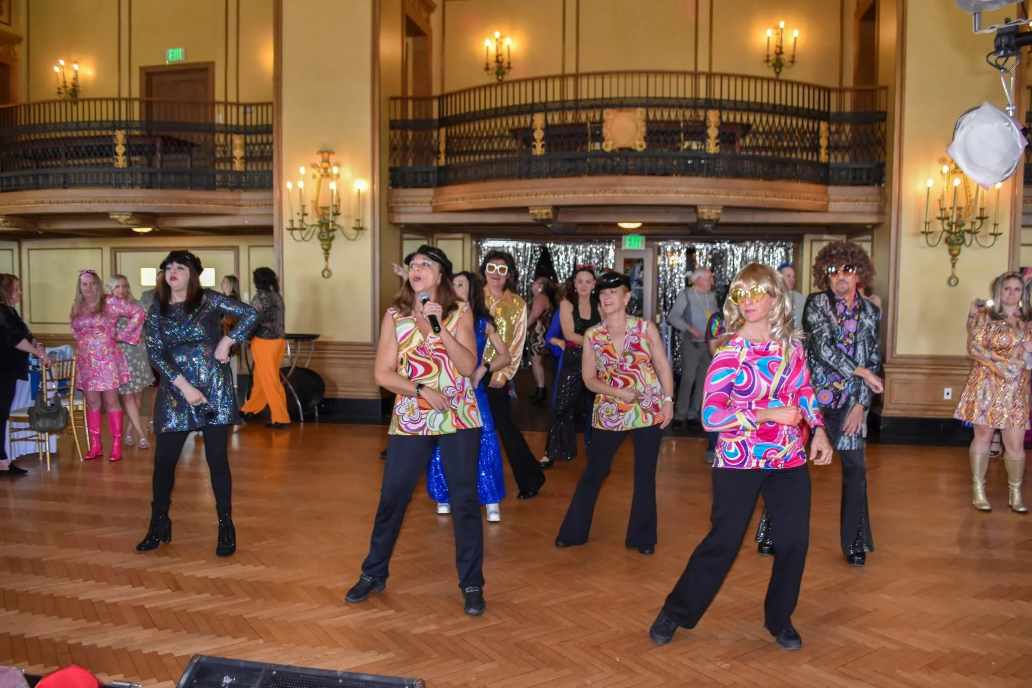 People dressed in colorful, retro-style clothing dancing on a wooden floor in a decorated banquet hall with chandeliers and an upper balcony.