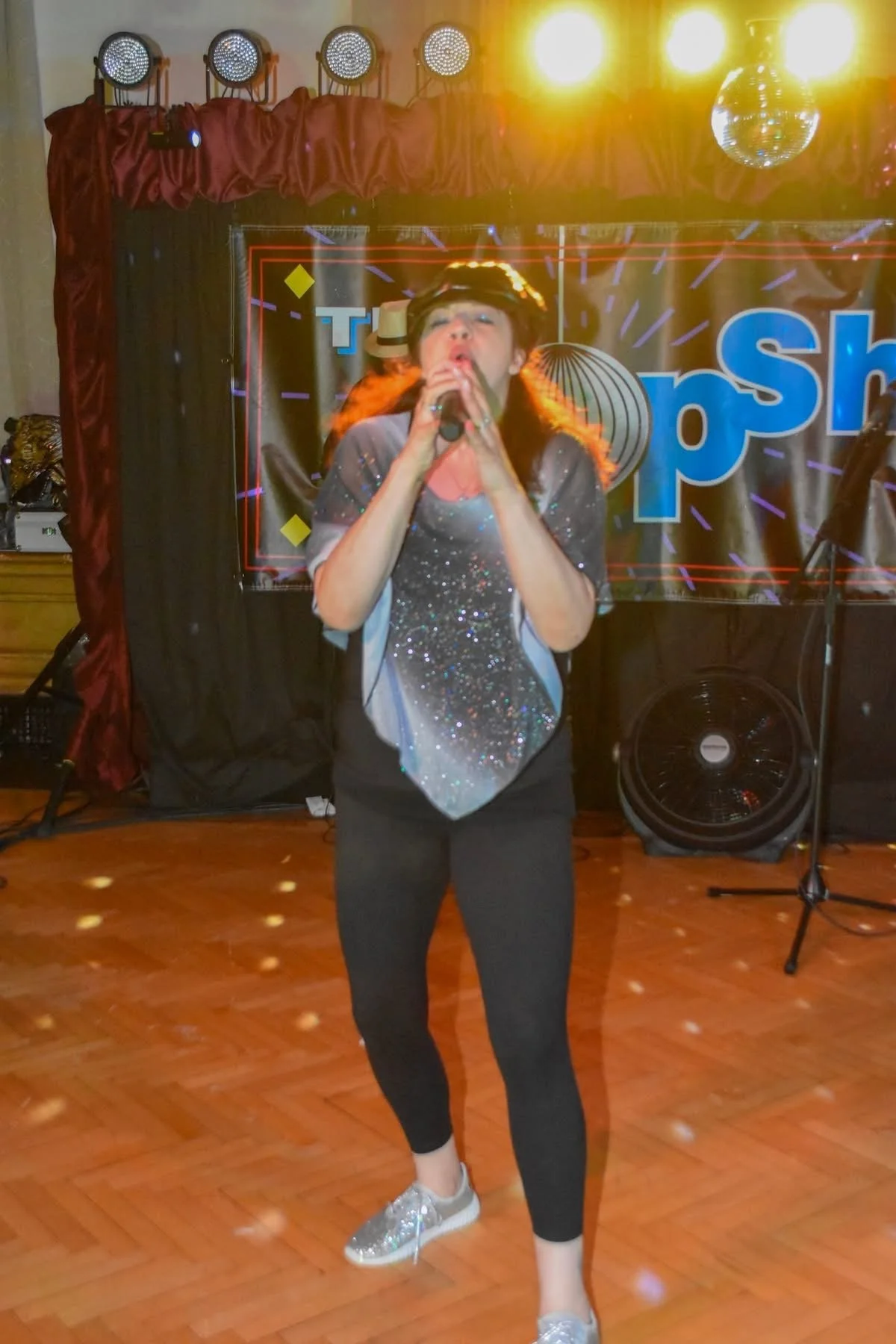 A woman singing into a microphone on stage in a club or event setting with a black backdrop, red curtains, and a disco ball overhead. She is wearing a sparkly top and black pants with silver shoes, with warm stage lighting.